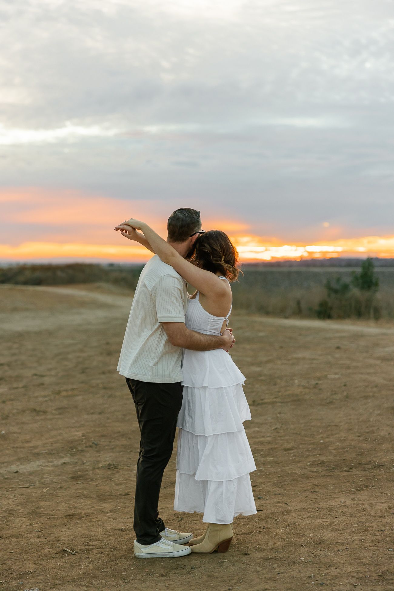 A couple embraces while watching a colorful sunset over a desert landscape.