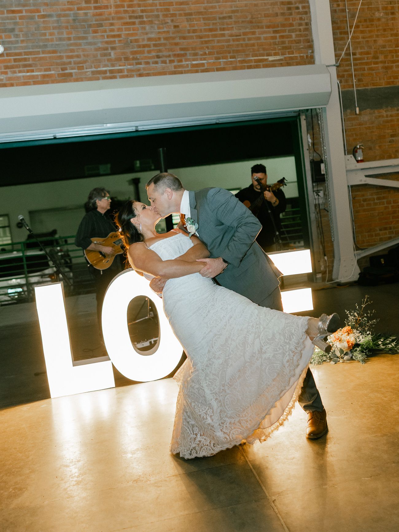 Couple performs romantic dip during first dance with illuminated letters and brick walls in background.