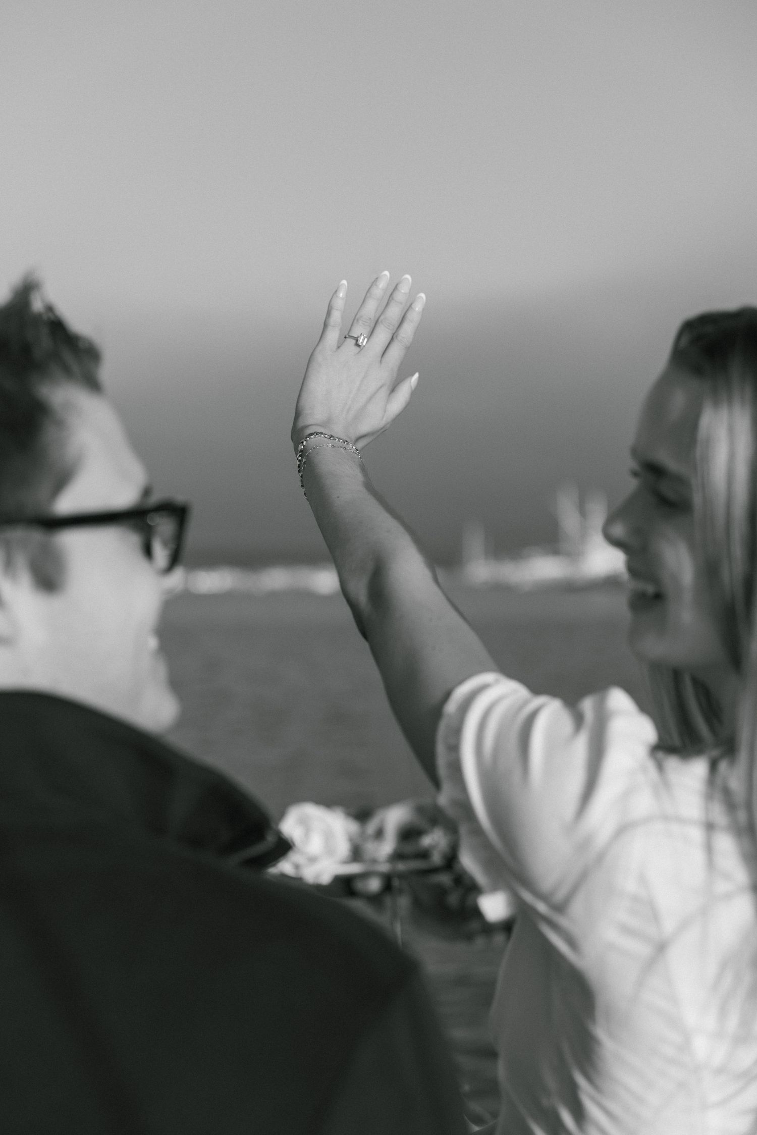 A black and white photo showing a woman's hand with an engagement ring raised against an ocean backdrop.