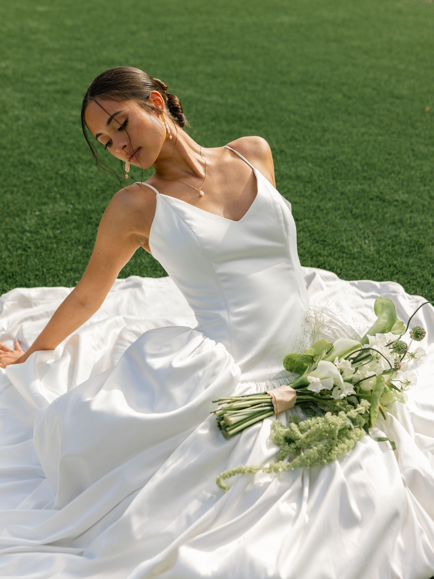 A flowing white wedding dress spreads across green grass with a delicate white floral bouquet.