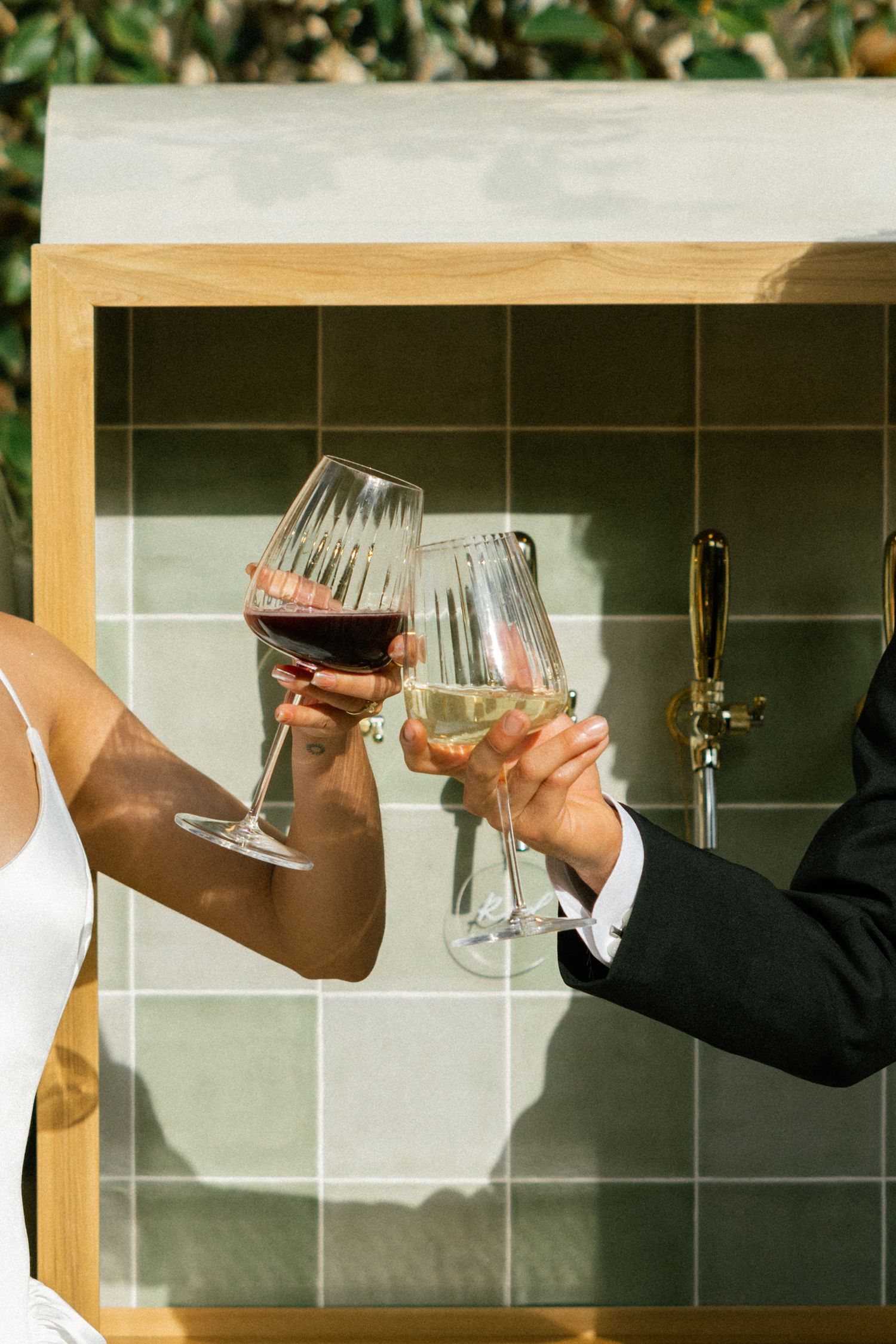 Two hands toast with wine glasses against a tiled backdrop at an outdoor celebration.