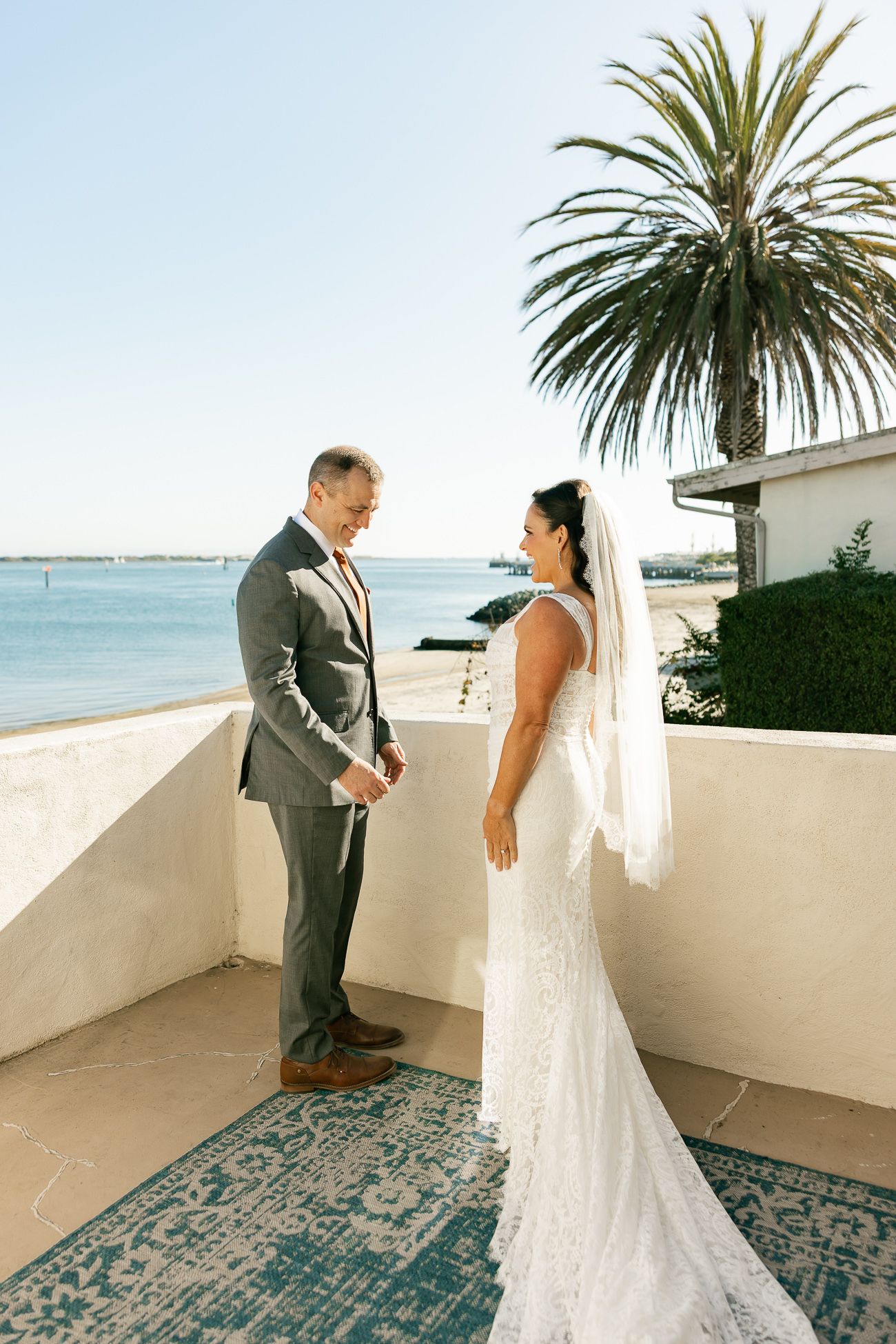 A couple shares a wedding moment on a coastal balcony with a palm tree and ocean view in the background.