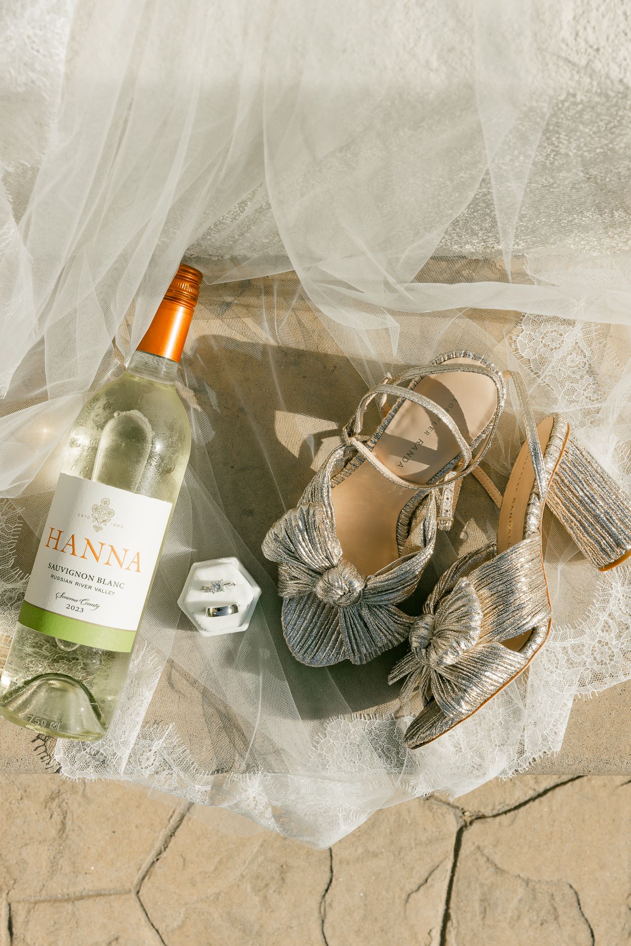 A bottle of white wine and metallic silver bridal shoes arranged on tulle fabric and stone flooring.