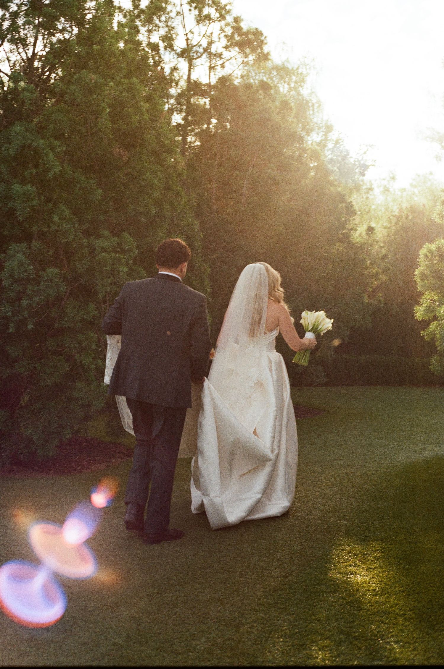 A bride and groom walk together on a sunlit lawn during golden hour, with lens flares visible in the foreground.