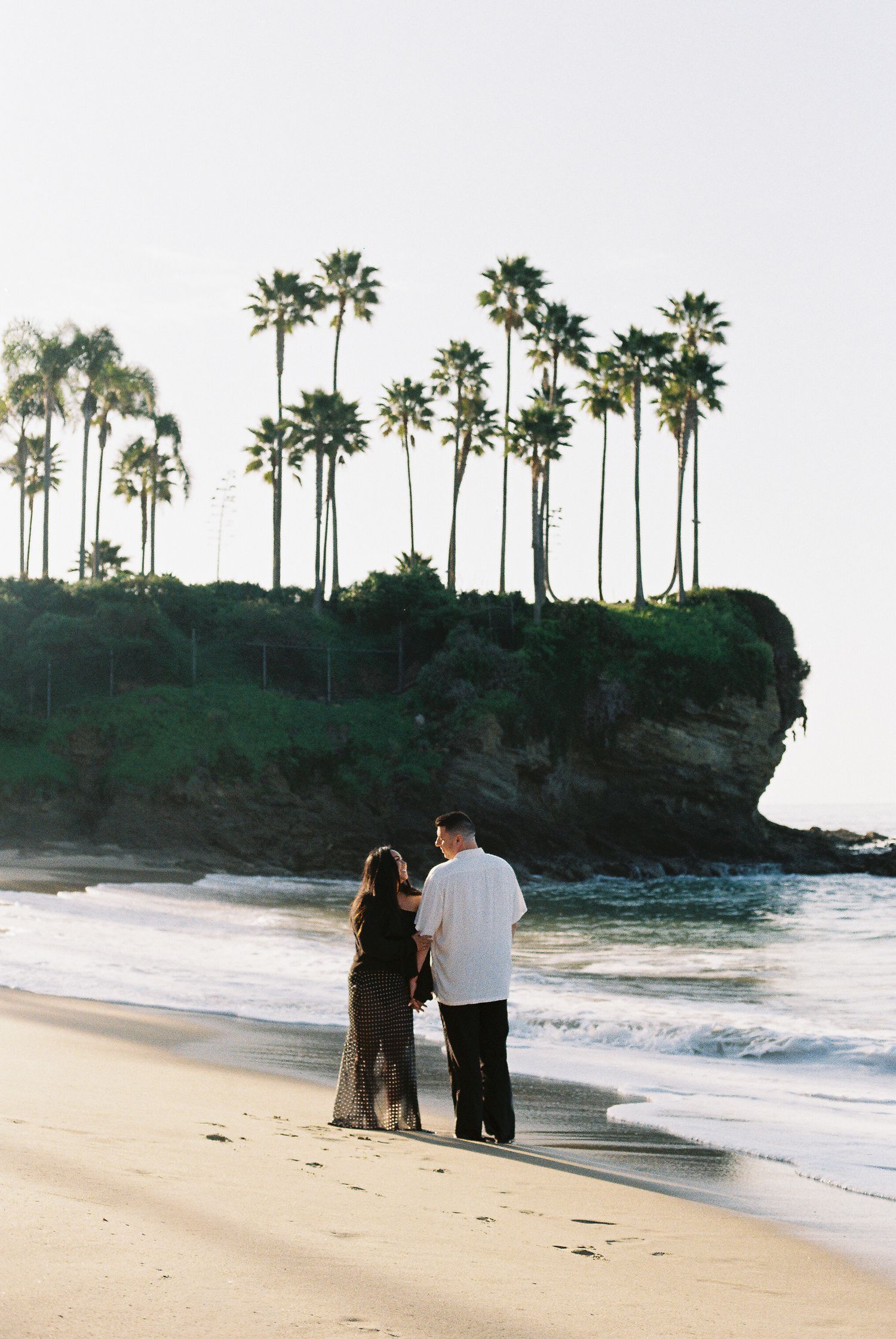 A couple stands on a scenic beach with tall palm trees on a cliff overlooking the ocean at sunset.