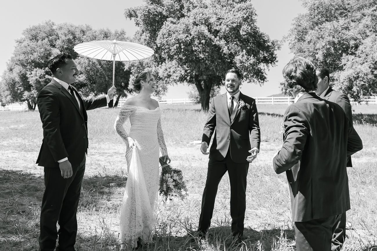 A black and white wedding photo showing a small group gathered outdoors with a white parasol and bouquet.