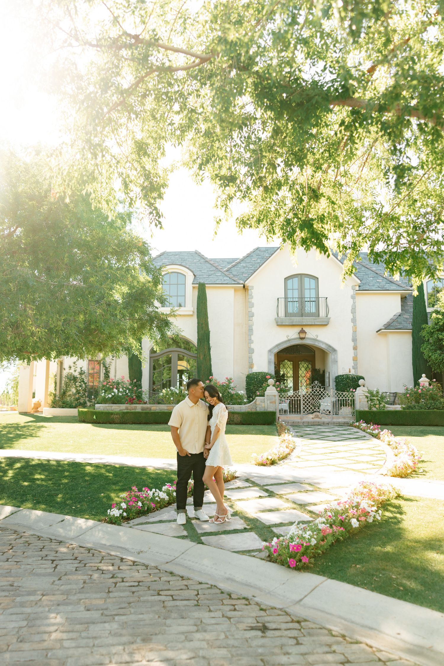 A stone pathway leads to a beautiful French country style home with landscaped gardens in bright sunlight.