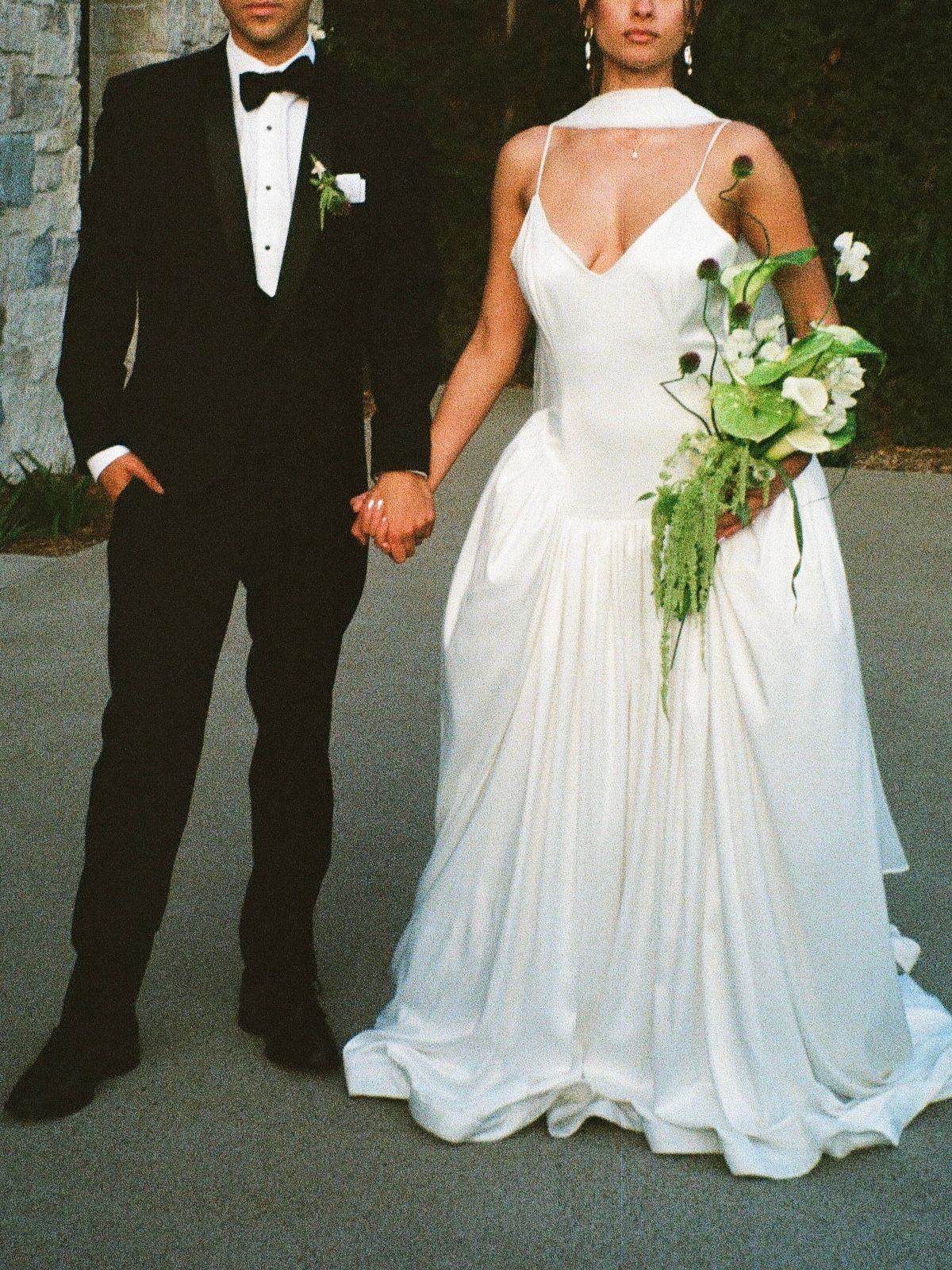 A couple in formal wedding attire with black tuxedo and white gown holding hands while carrying a cascading white bouquet.