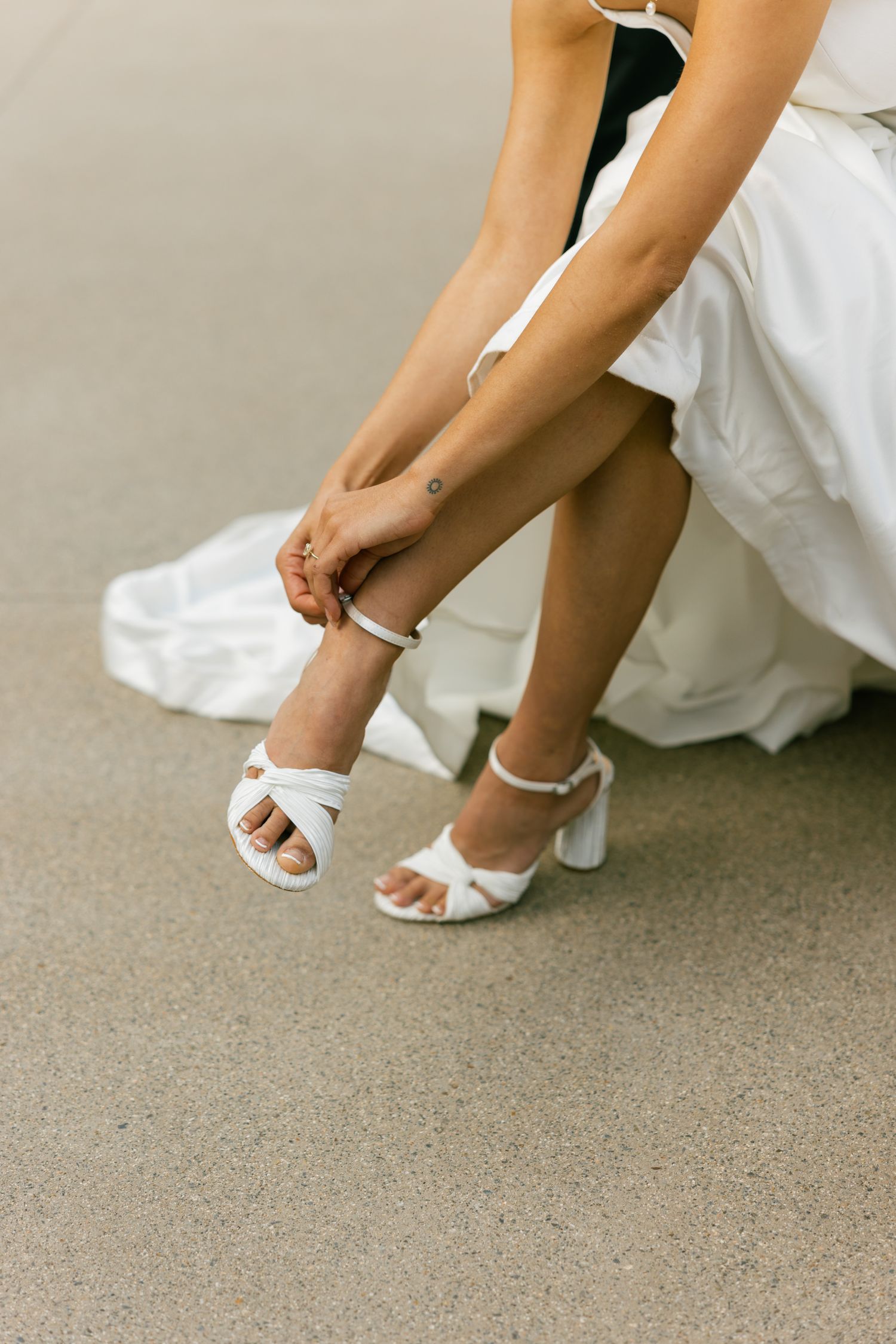 A person wearing a white dress adjusts the strap on elegant white block heel sandals while seated.