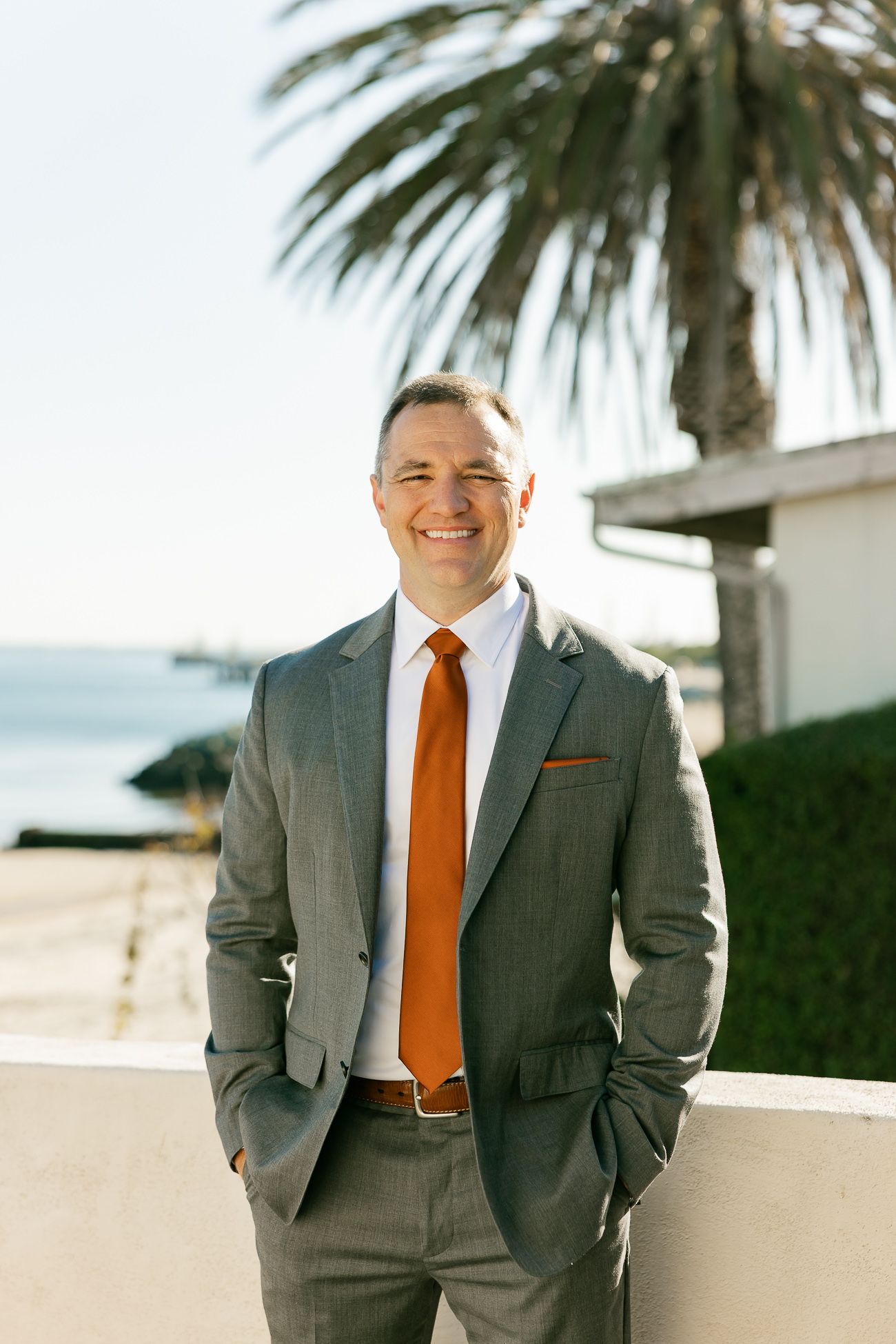 A professional stands against a coastal backdrop wearing a grey suit and orange tie with a palm tree visible above.