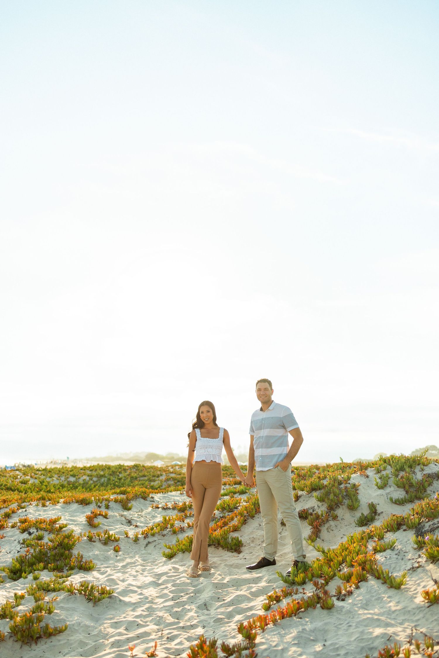 Two people walk together on sandy beach dunes with scattered vegetation at sunset.