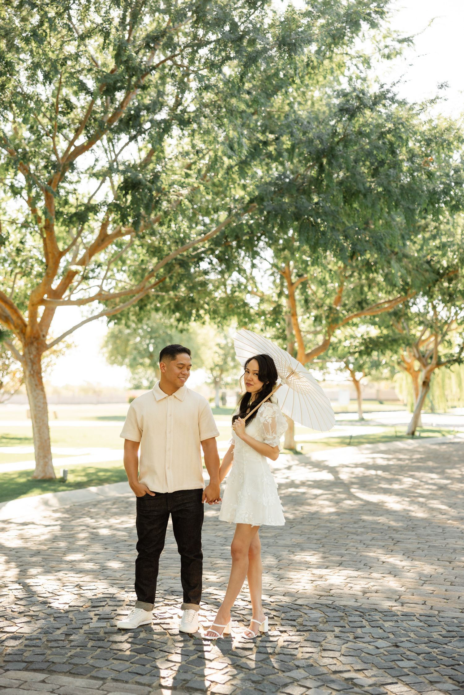 A romantic couple shares playful moments together under sunlit trees in a park setting.