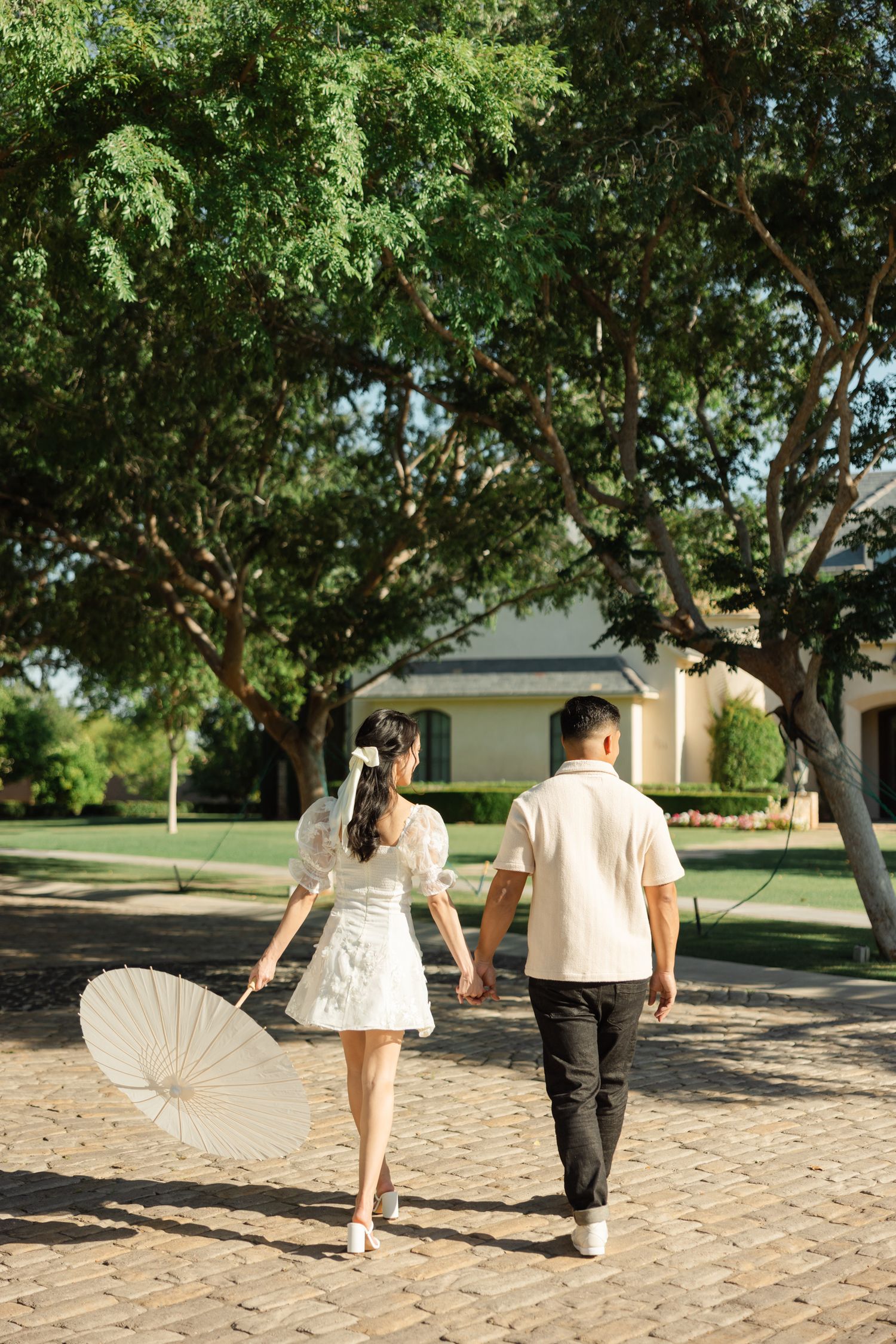 A couple walks hand in hand down a brick path under shady trees while holding a white parasol.