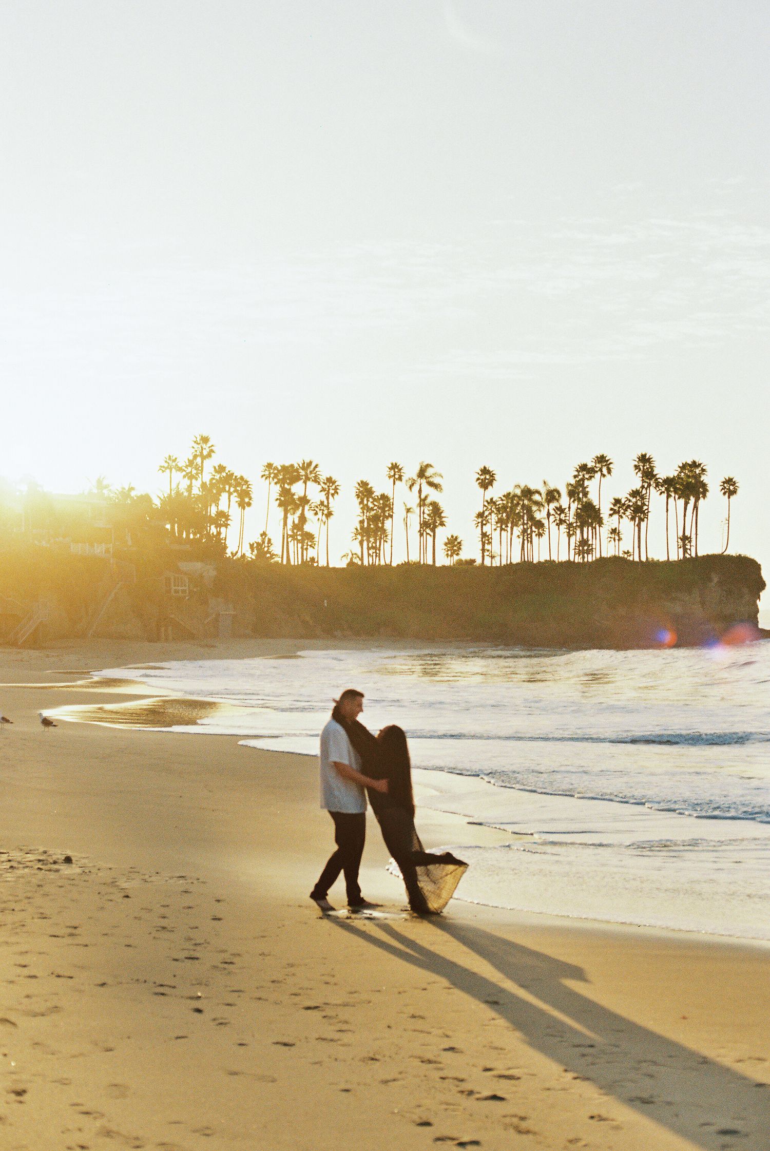 Couple walking on a beach at sunset with palm trees silhouetted on the cliffside in the background.