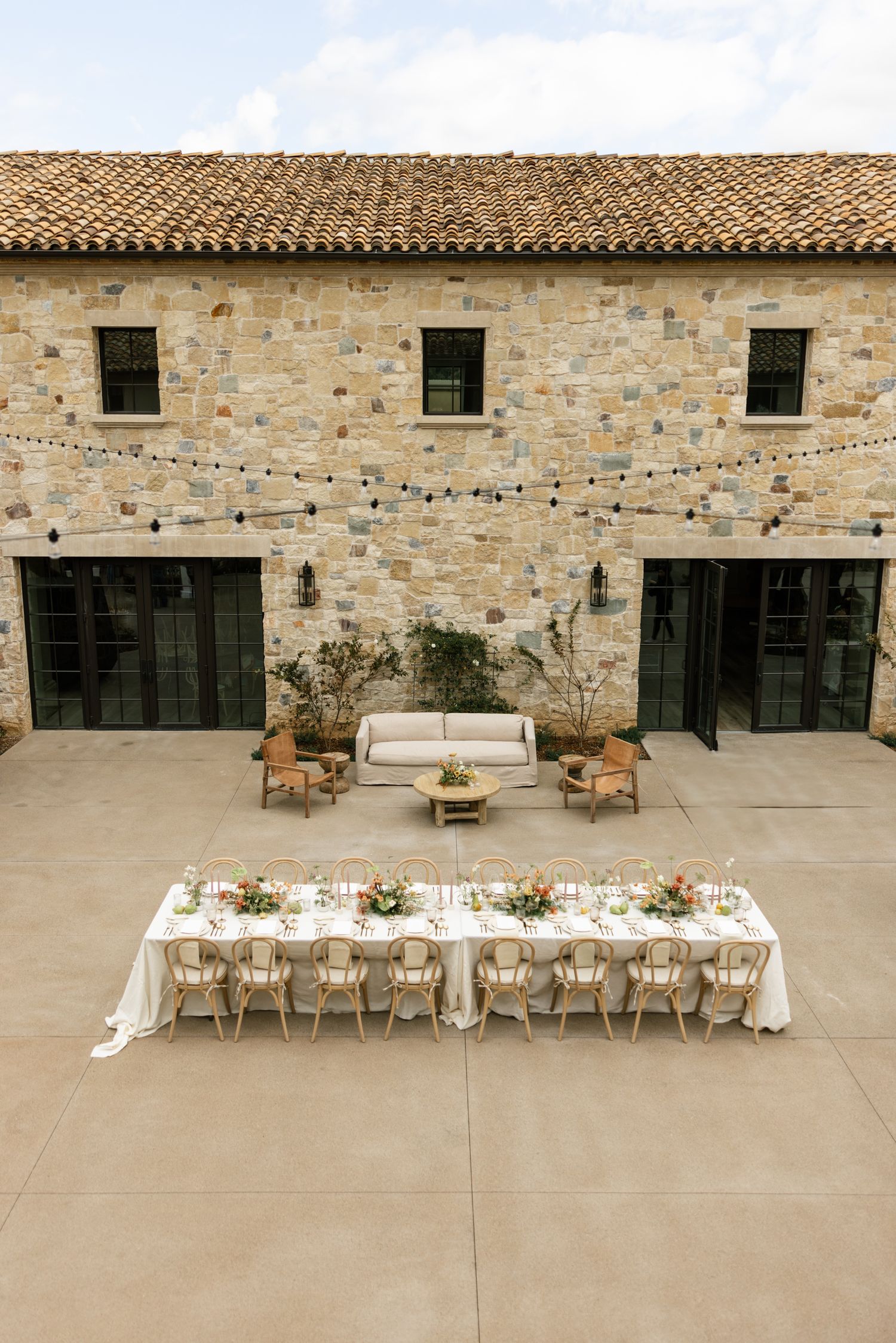 Outdoor wedding reception setup with long white table against rustic stone building facade.