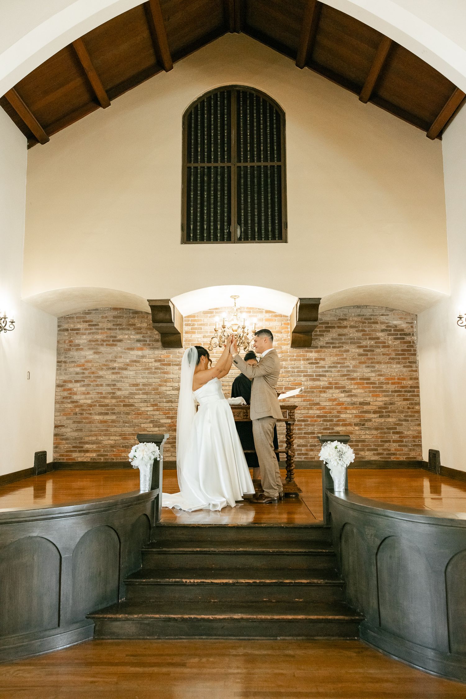 A couple shares their first kiss at a rustic chapel altar with brick accents and wooden beams overhead.