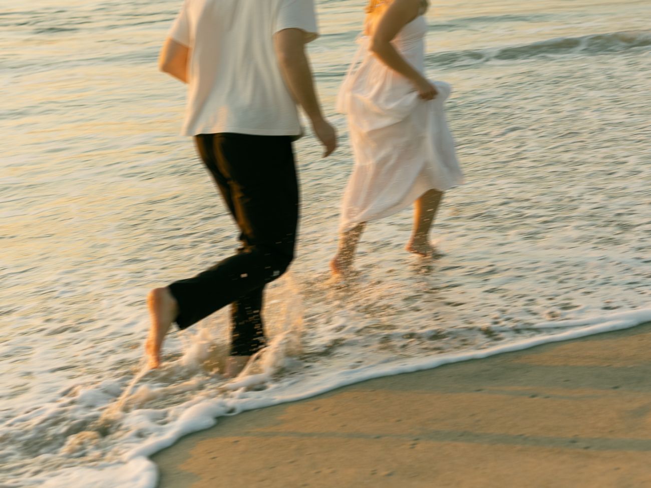 Two people walking barefoot along the beach shoreline at sunset with waves rolling in.