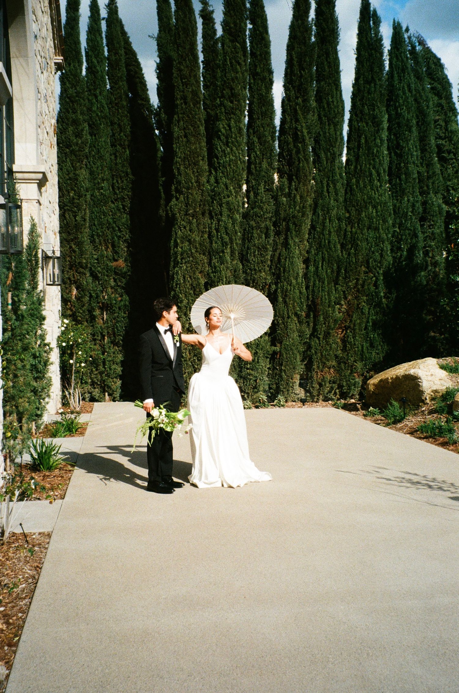 Elegant wedding scene with cypress trees lining a garden path while couple poses with white parasol.