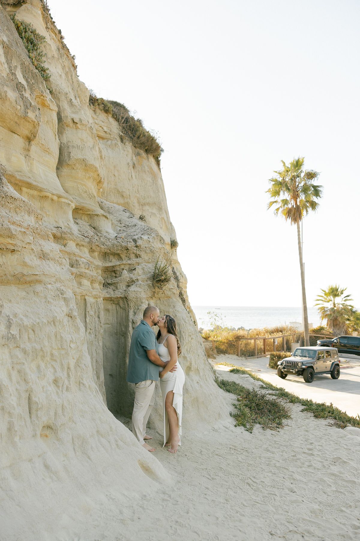 A romantic beach photoshoot sequence showing a couple embracing near coastal cliffs at sunset with palm trees in view.