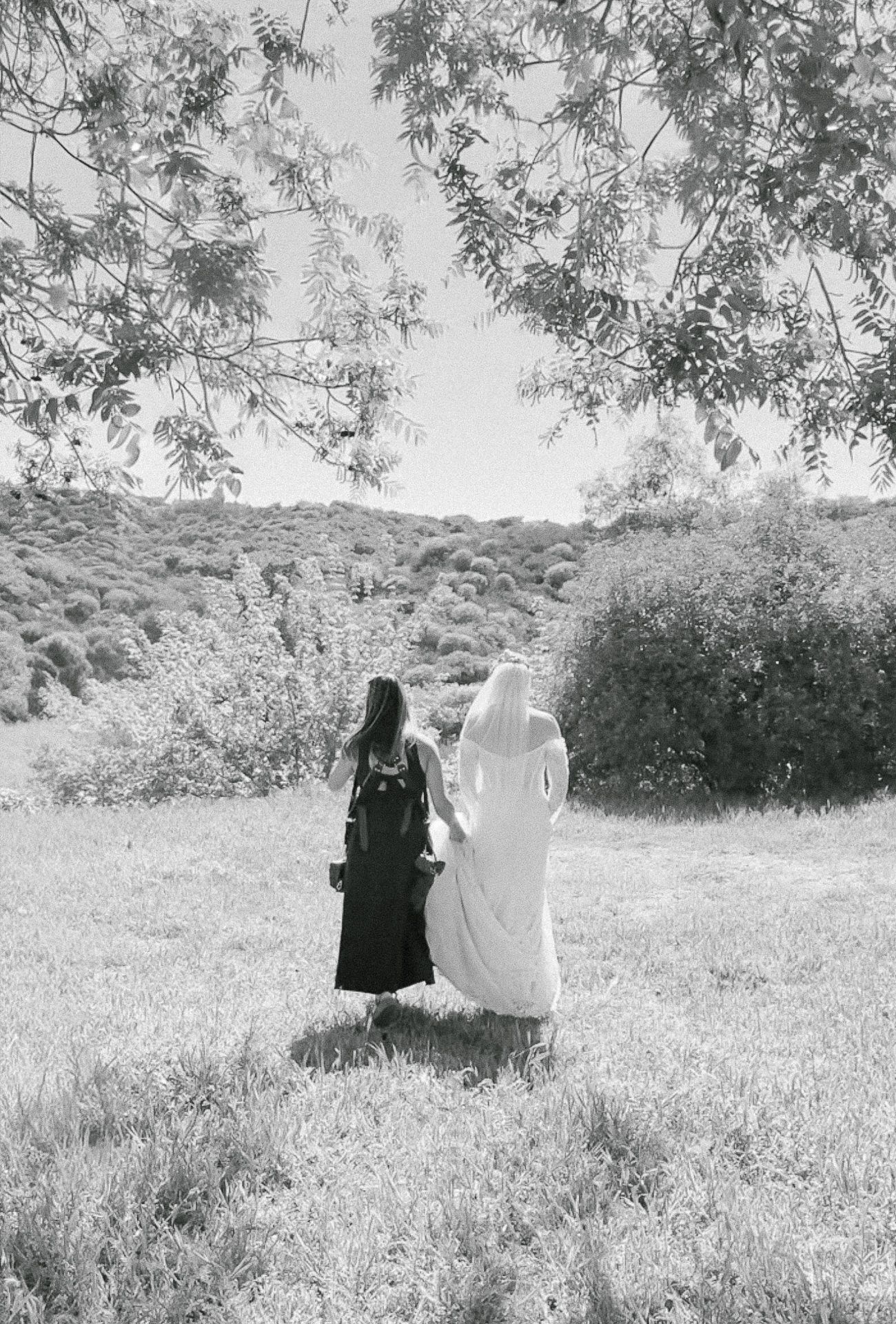 Two figures in contrasting black and white outfits walk through a sunlit meadow in an artistic black and white photograph.