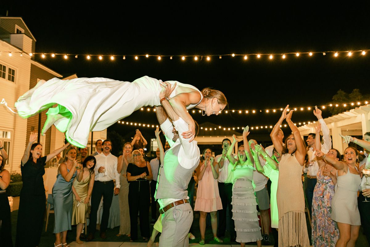 Wedding reception guests celebrate as a couple performs a dramatic lift under string lights.