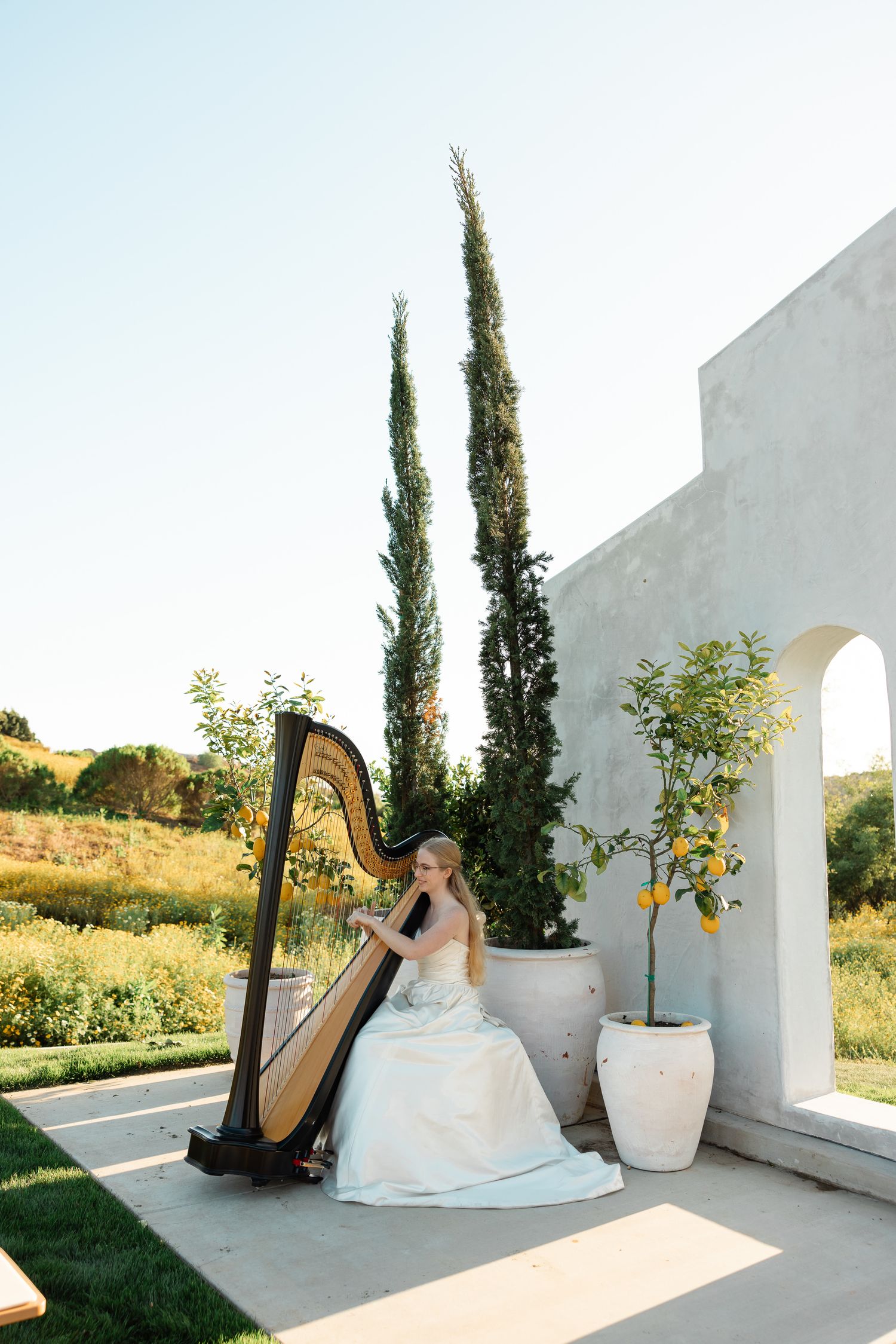 Musician in a white gown plays an elegant harp outdoors next to tall cypress trees and white architecture.