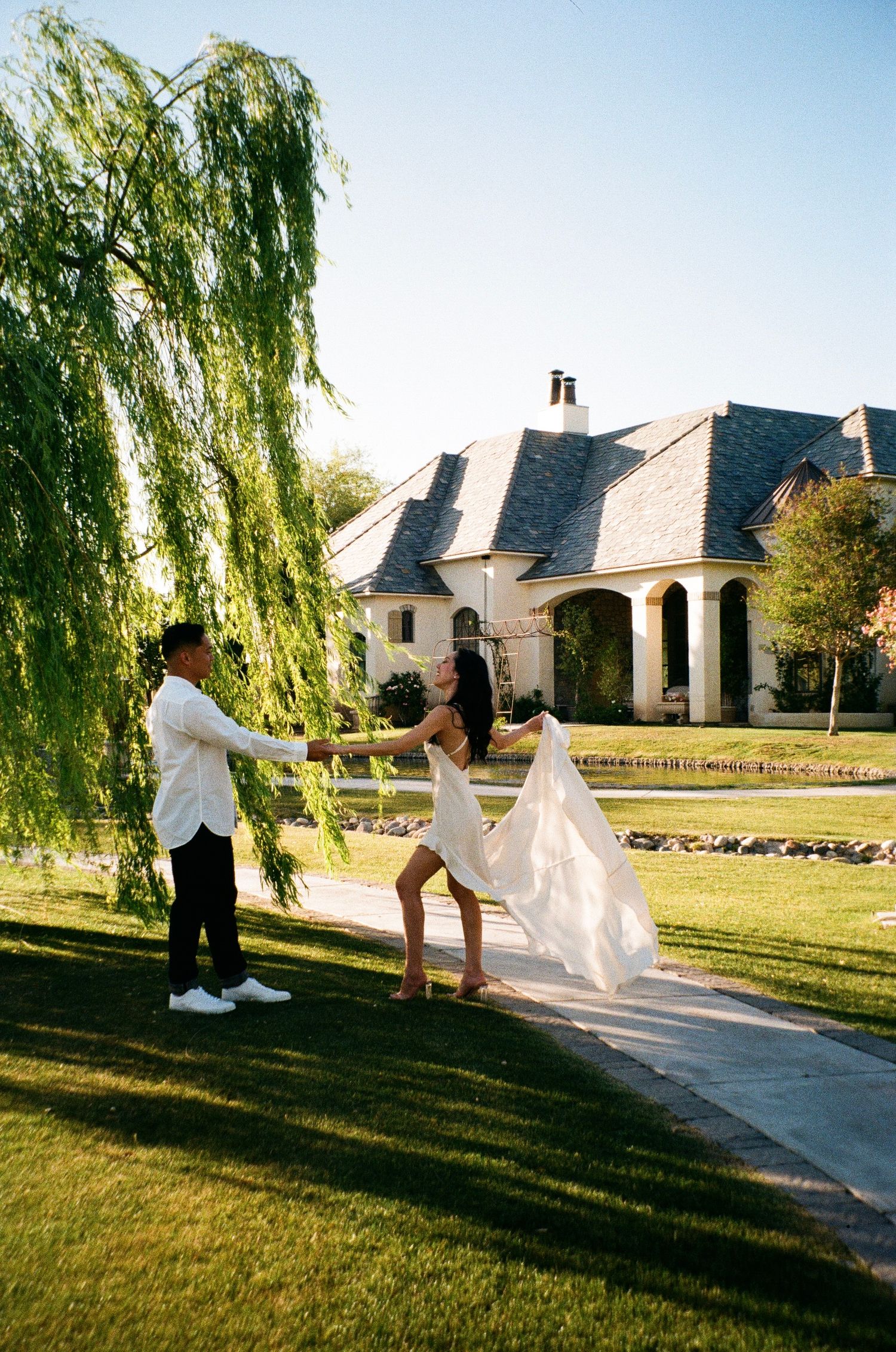 A joyful moment captured on a lawn with a weeping willow tree and elegant mansion in the background.