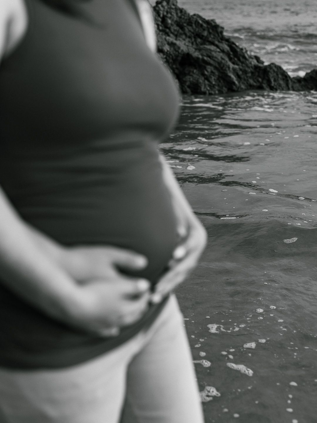A black and white close-up photo of a pregnant woman's belly at the beach with waves in the background.