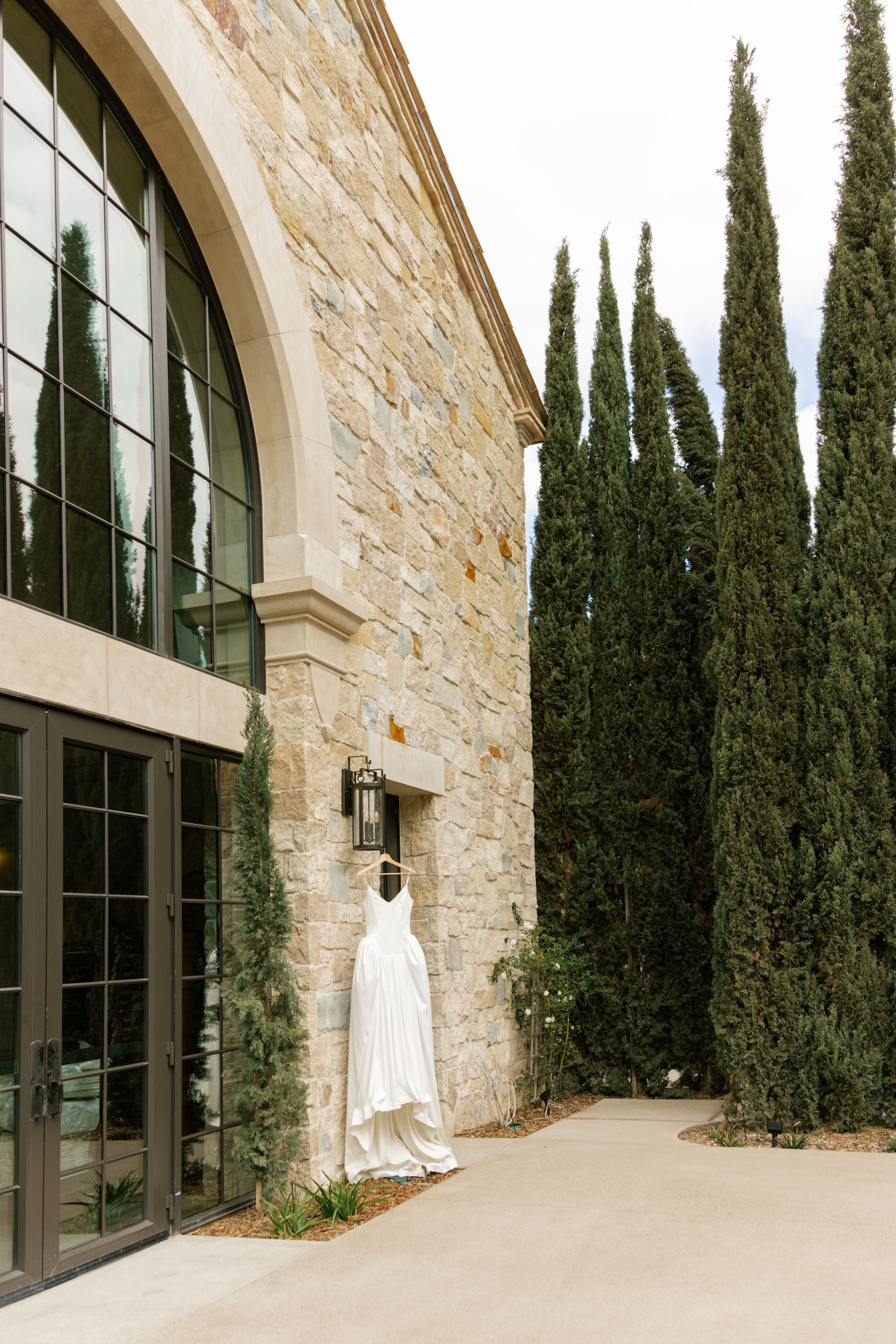 A white wedding dress hangs outside a stone building with arched windows and tall cypress trees in the background.