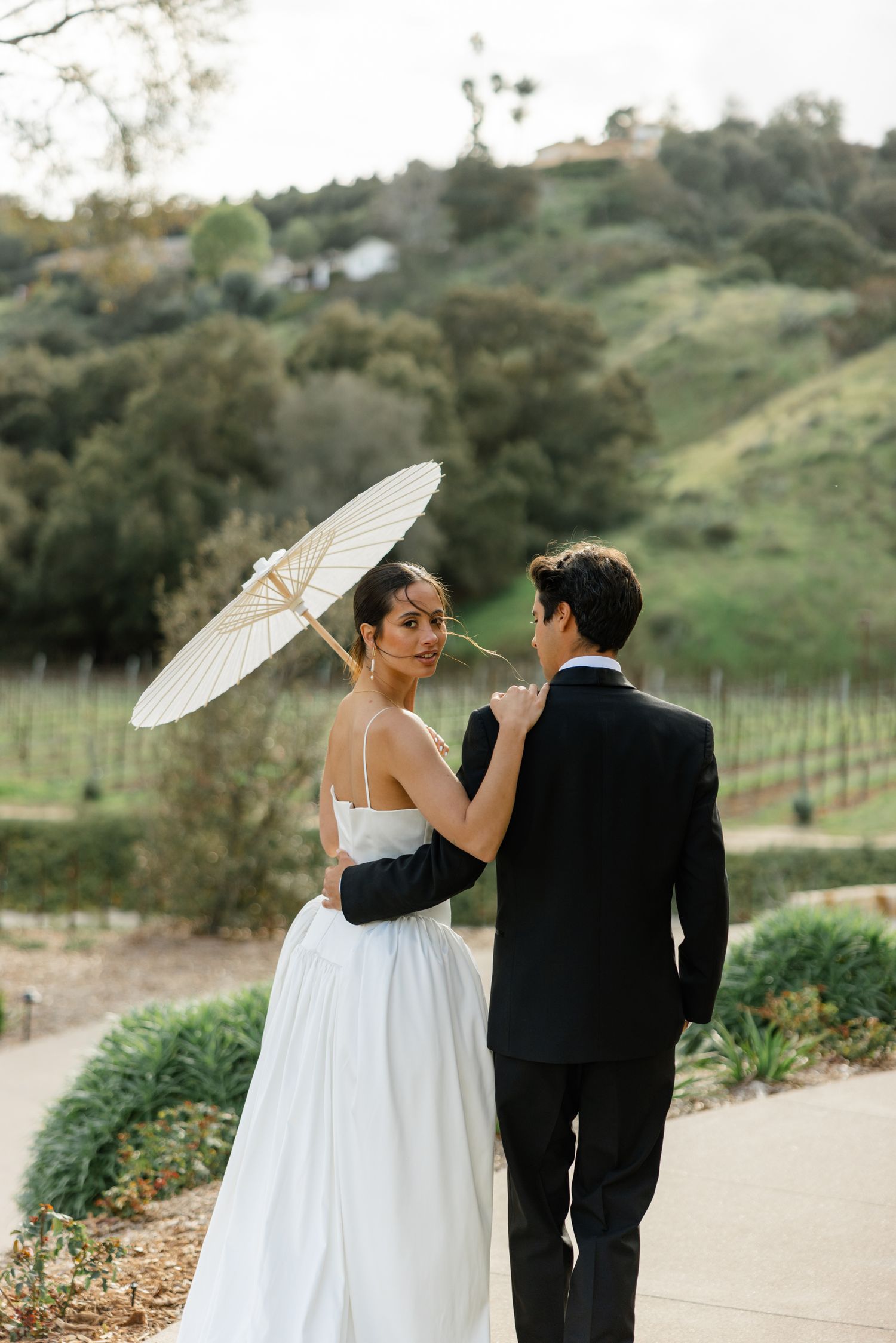 A romantic wedding couple poses on a garden path with a white parasol against rolling vineyard hills.