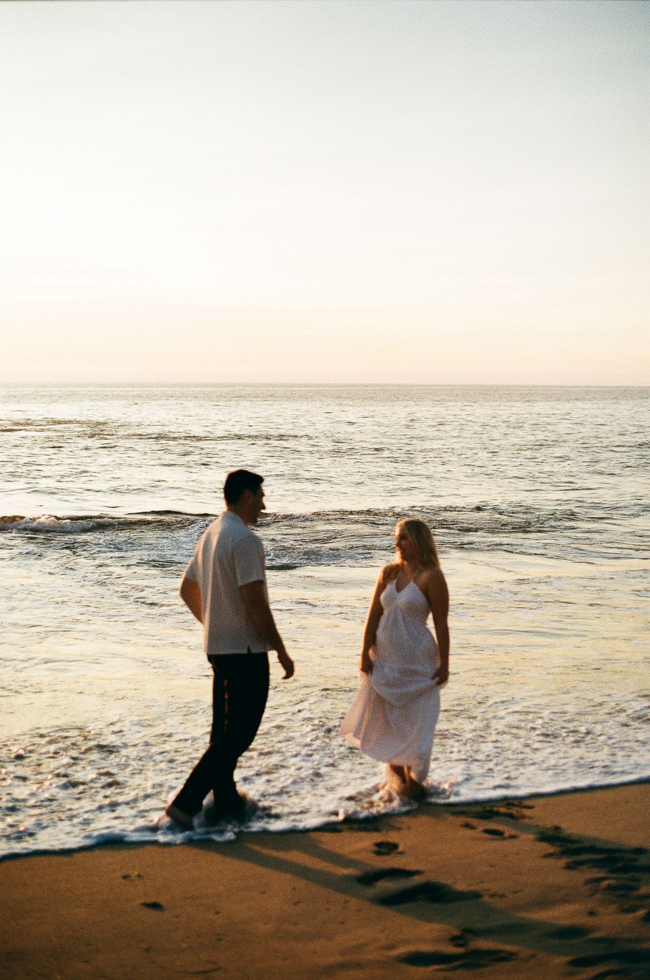 Two people walking together along a sandy beach at sunset with gentle waves washing up on the shore.