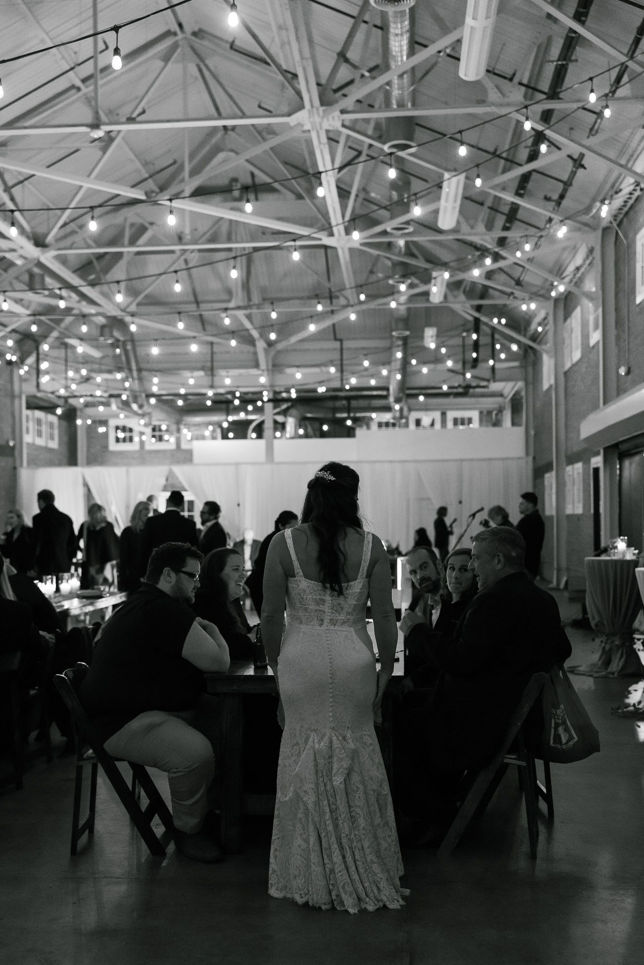 A black and white photo of a wedding reception in an industrial venue with string lights and guests seated at tables.