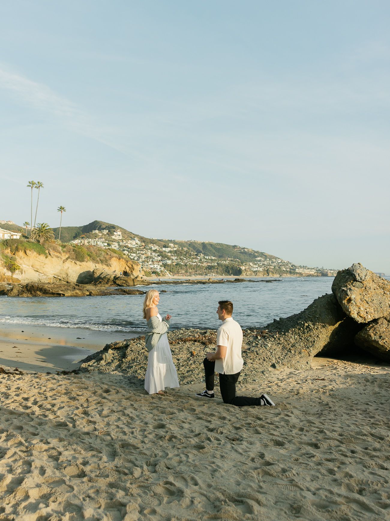A proposal scene unfolds on a rocky beach with coastal homes and palm trees visible in the background.