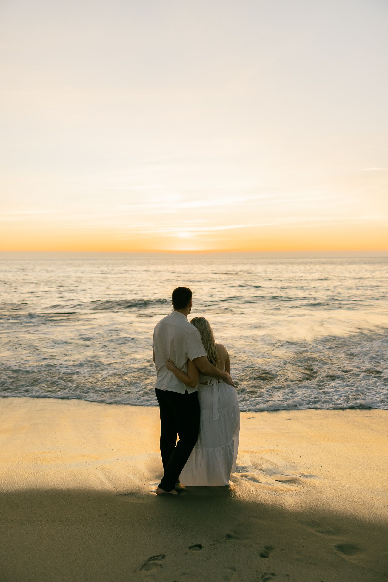 A couple embraces while watching the sunset over the ocean on a sandy beach with footprints in the sand.