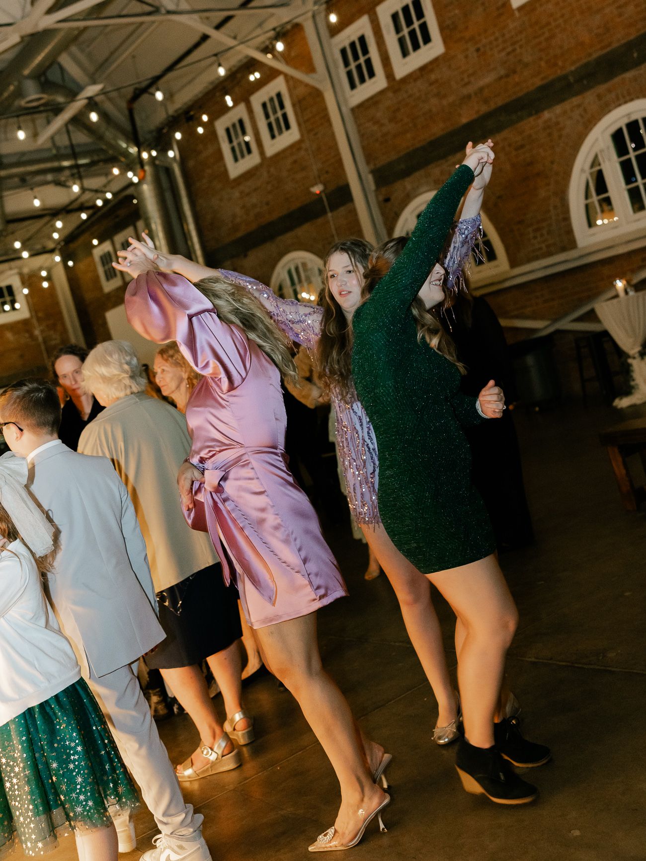 Wedding guests dance energetically under string lights in an industrial brick venue with high ceilings.