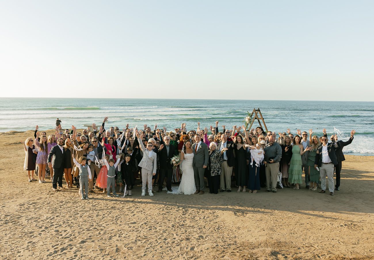 Large wedding party gathered on a sunny beach with waves and ocean in the background.