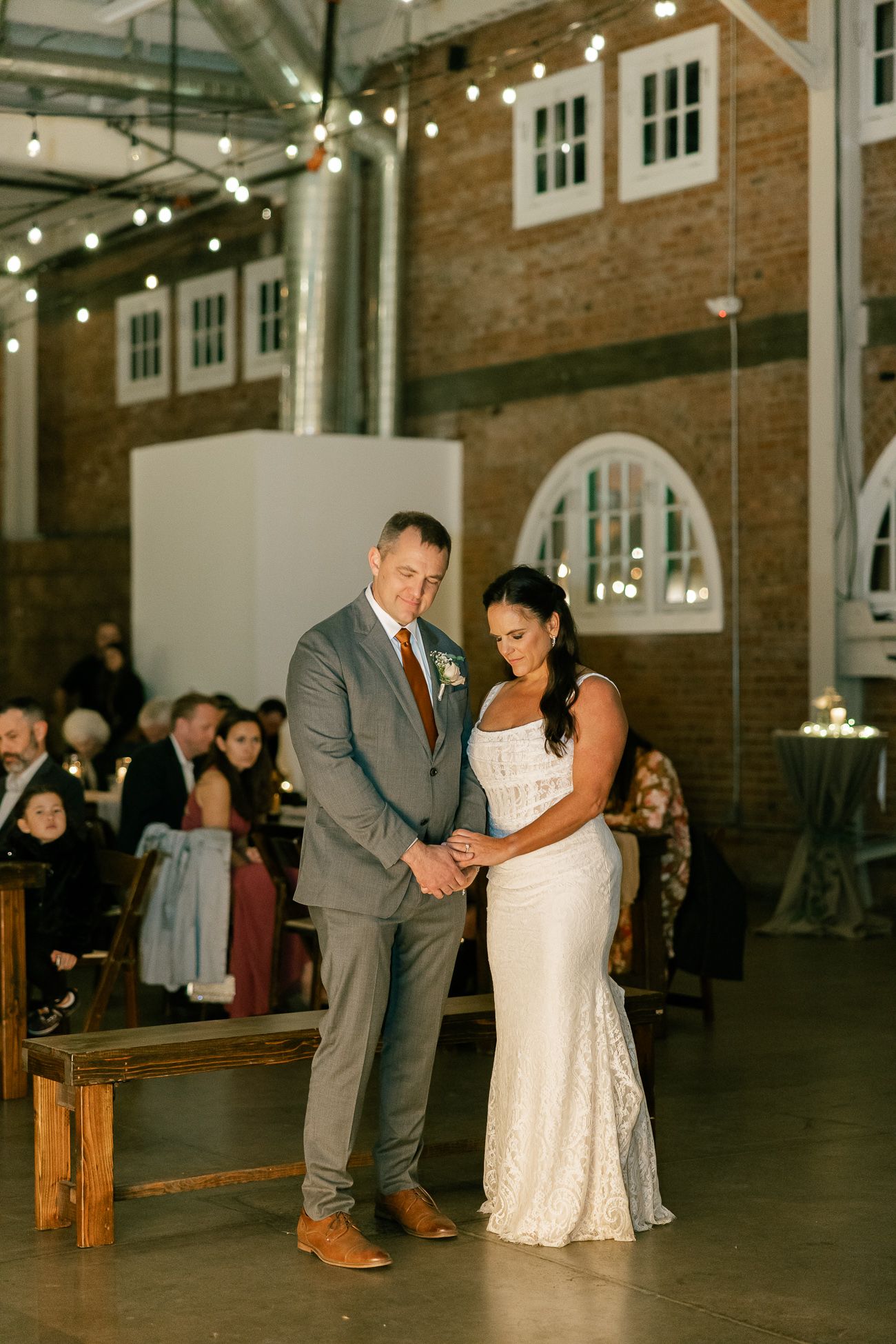 Wedding couple sharing an intimate moment in an industrial venue with string lights and exposed brick walls.