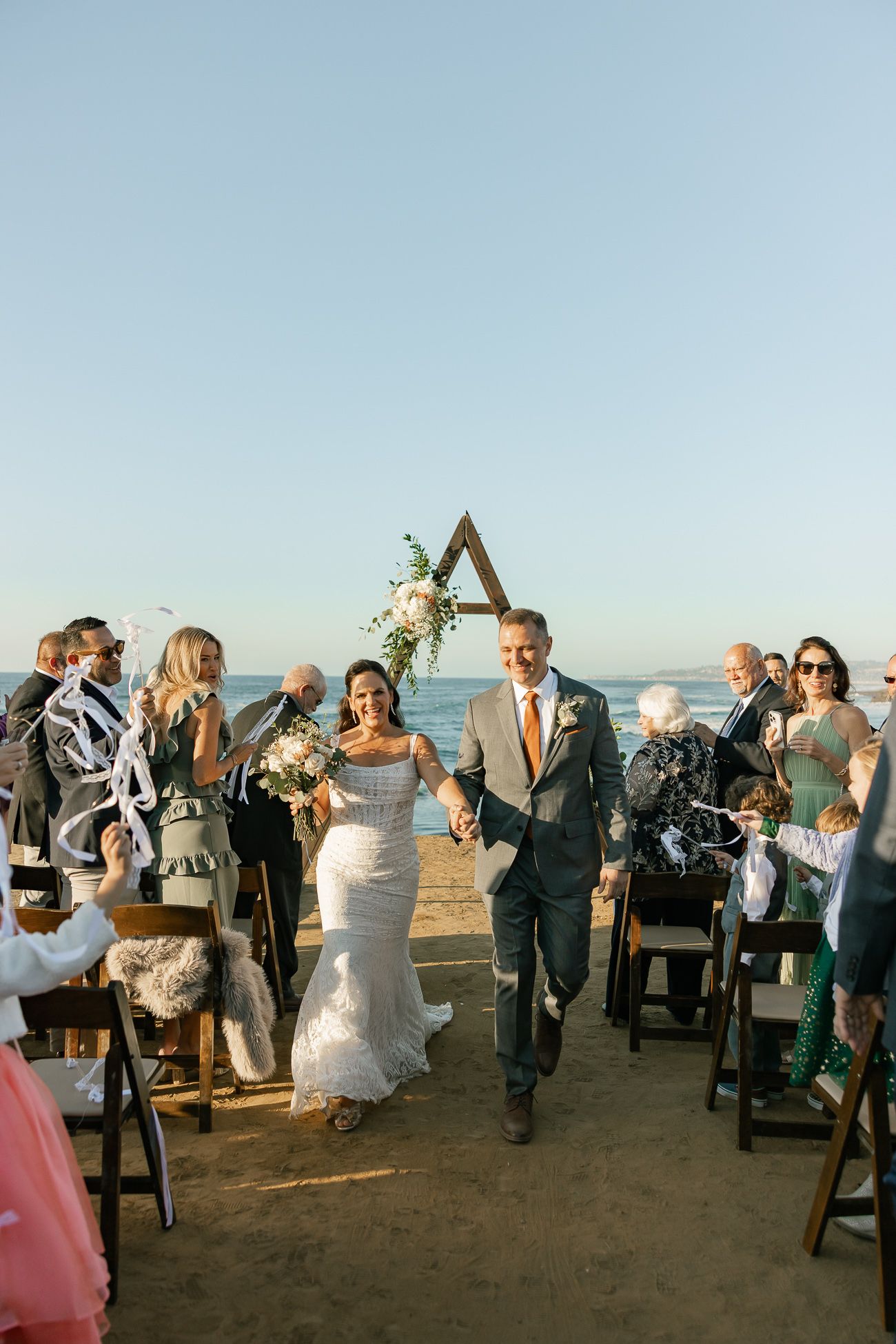 Wedding ceremony on beach with wooden triangle arch and seated guests as couple walks down aisle at sunset.