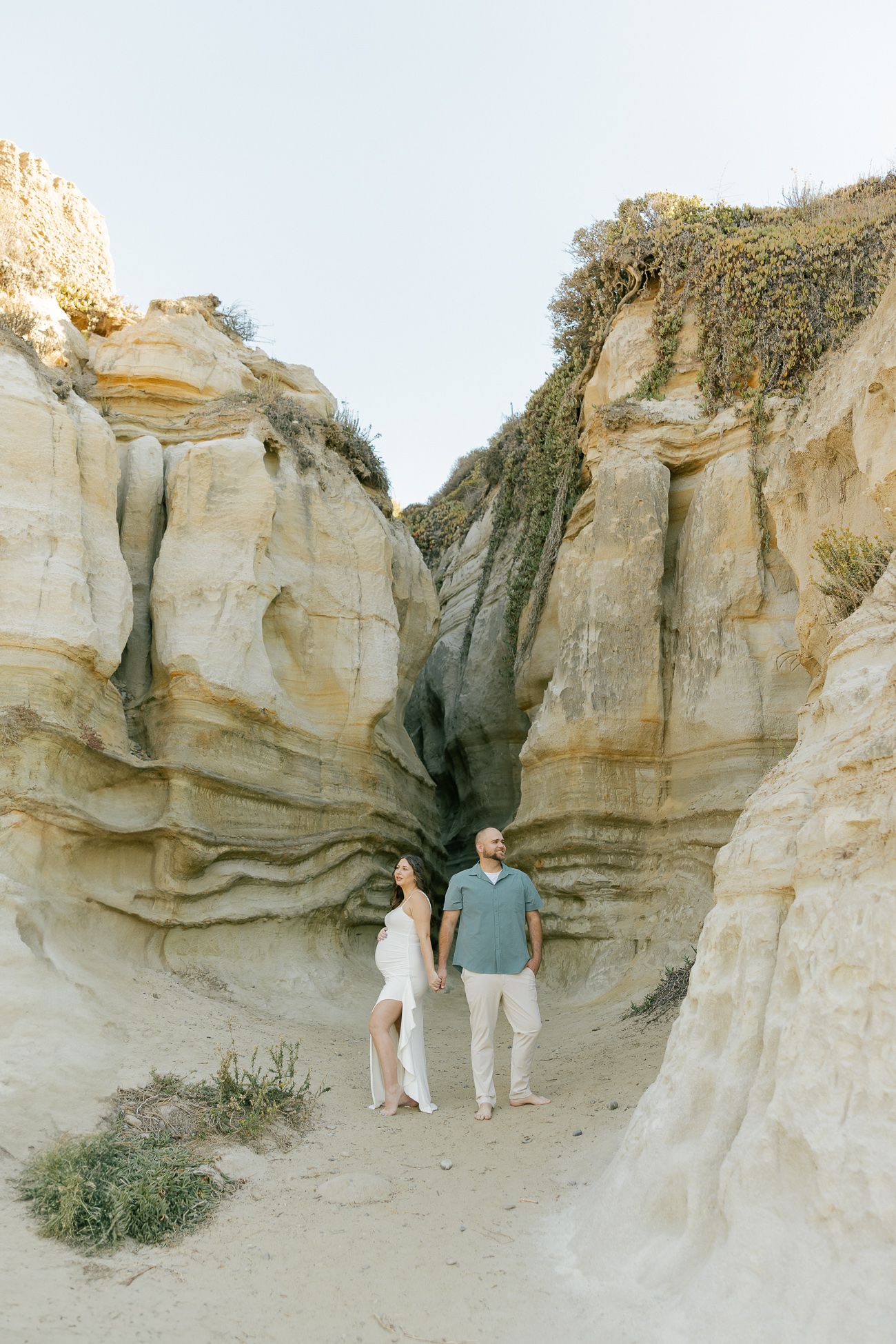 A panoramic view of desert rock formations with two people walking along the sandy path between towering limestone cliffs.
