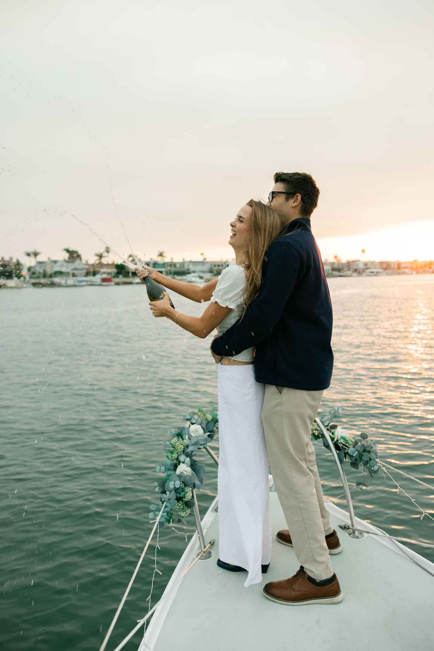 Two people share a romantic moment on the bow of a boat during sunset with coastal homes visible in the background.