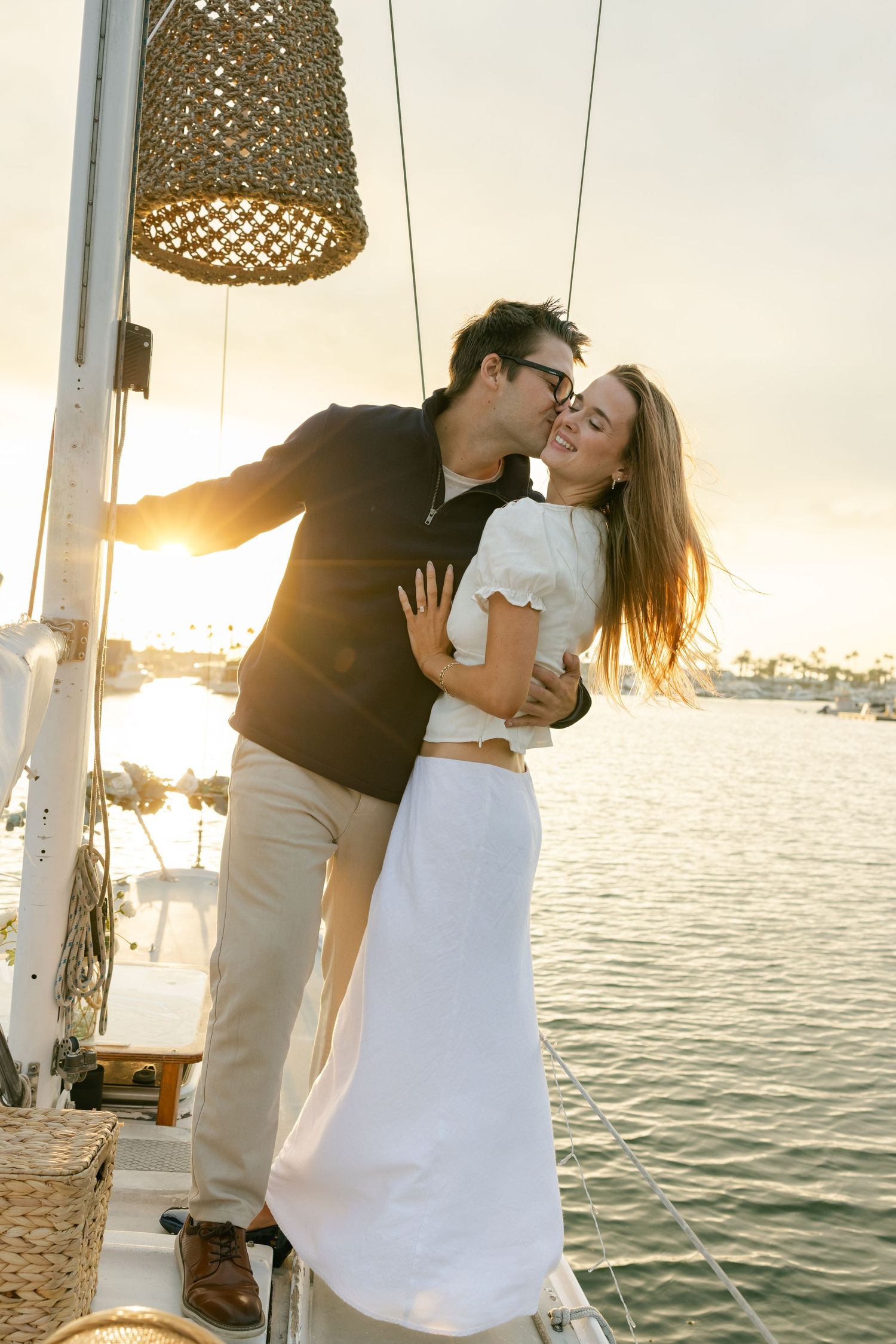 A romantic sunset moment aboard a sailboat with a woven lamp overhead and calm waters in the background.