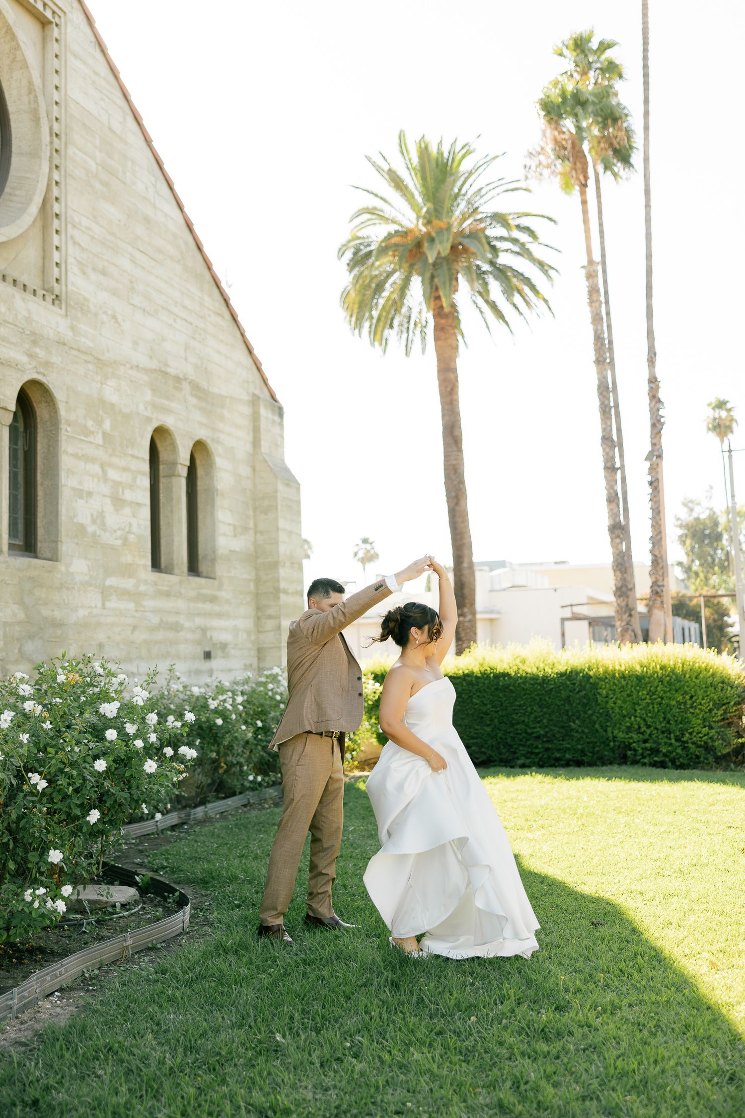 A joyful dance moment outside a stone church with blooming rose bushes and palm trees.