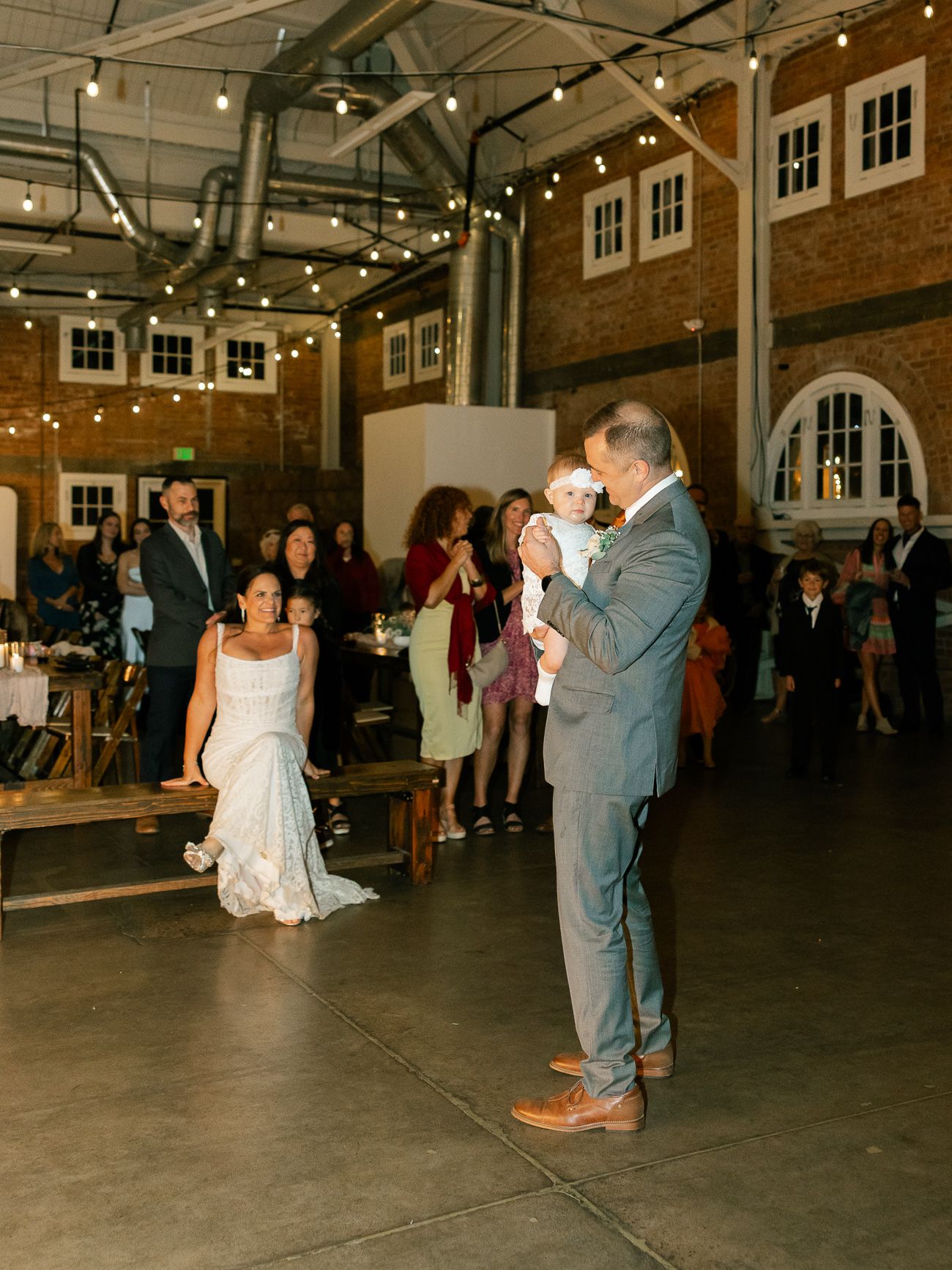 Couple dancing at a rustic indoor wedding reception with string lights overhead in a brick building venue.