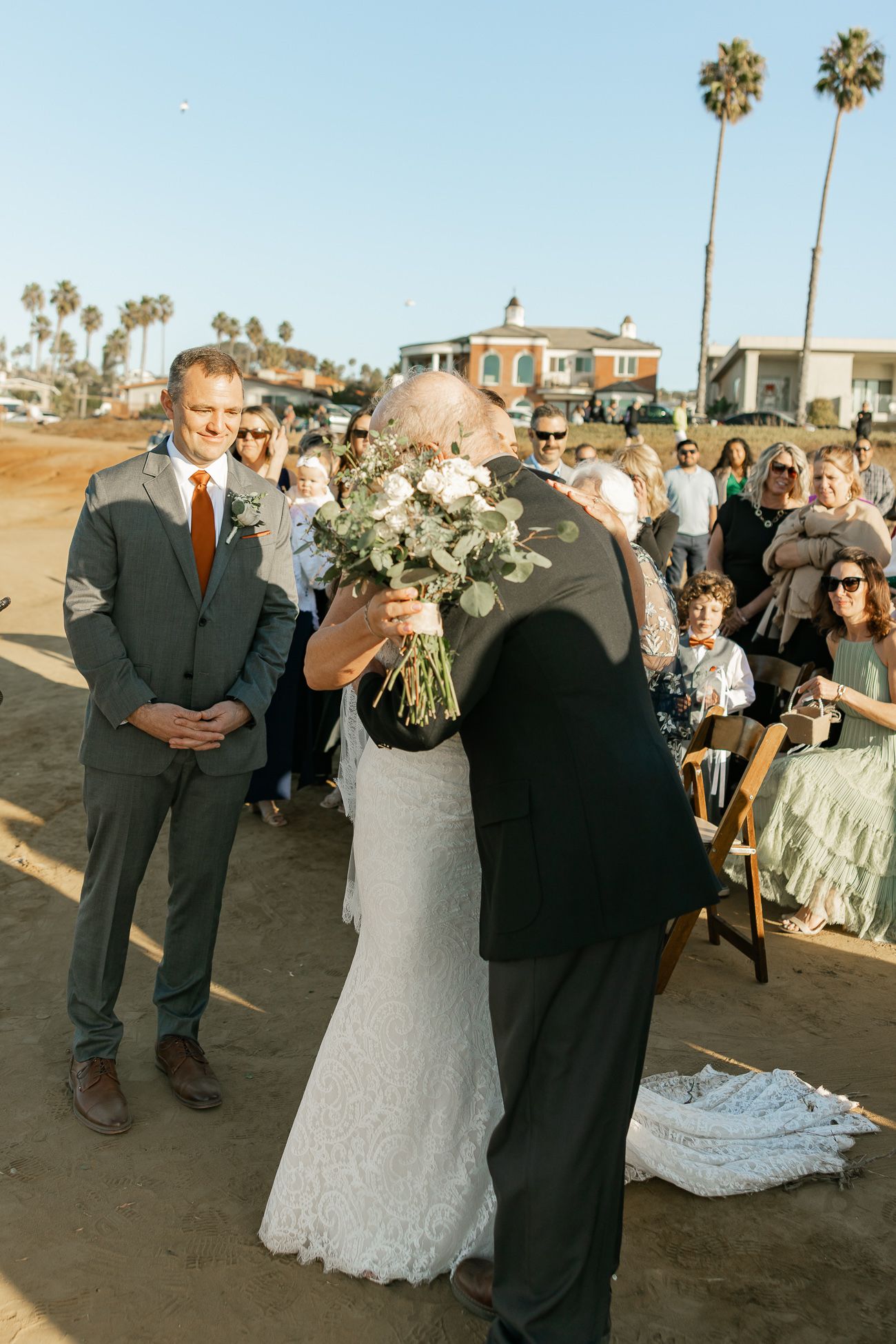 A beach wedding ceremony takes place with palm trees and guests gathered on the sand at sunset.