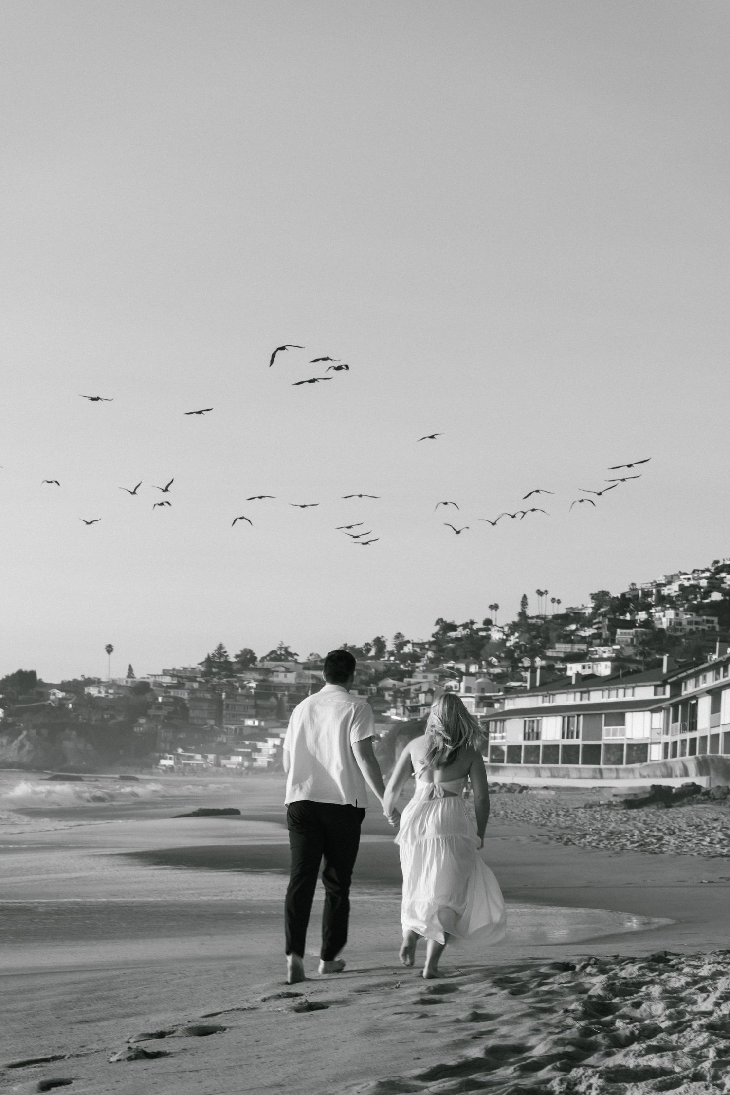 Couple walks hand in hand on beach with seabirds flying overhead in black and white photo.