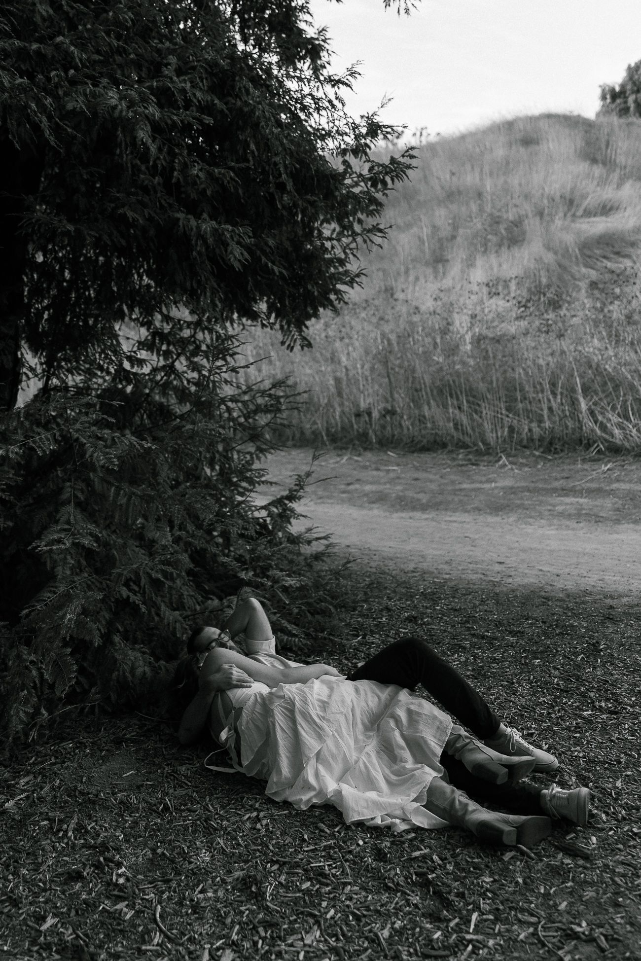 A black and white photo of two people lying on the ground beneath evergreen trees.