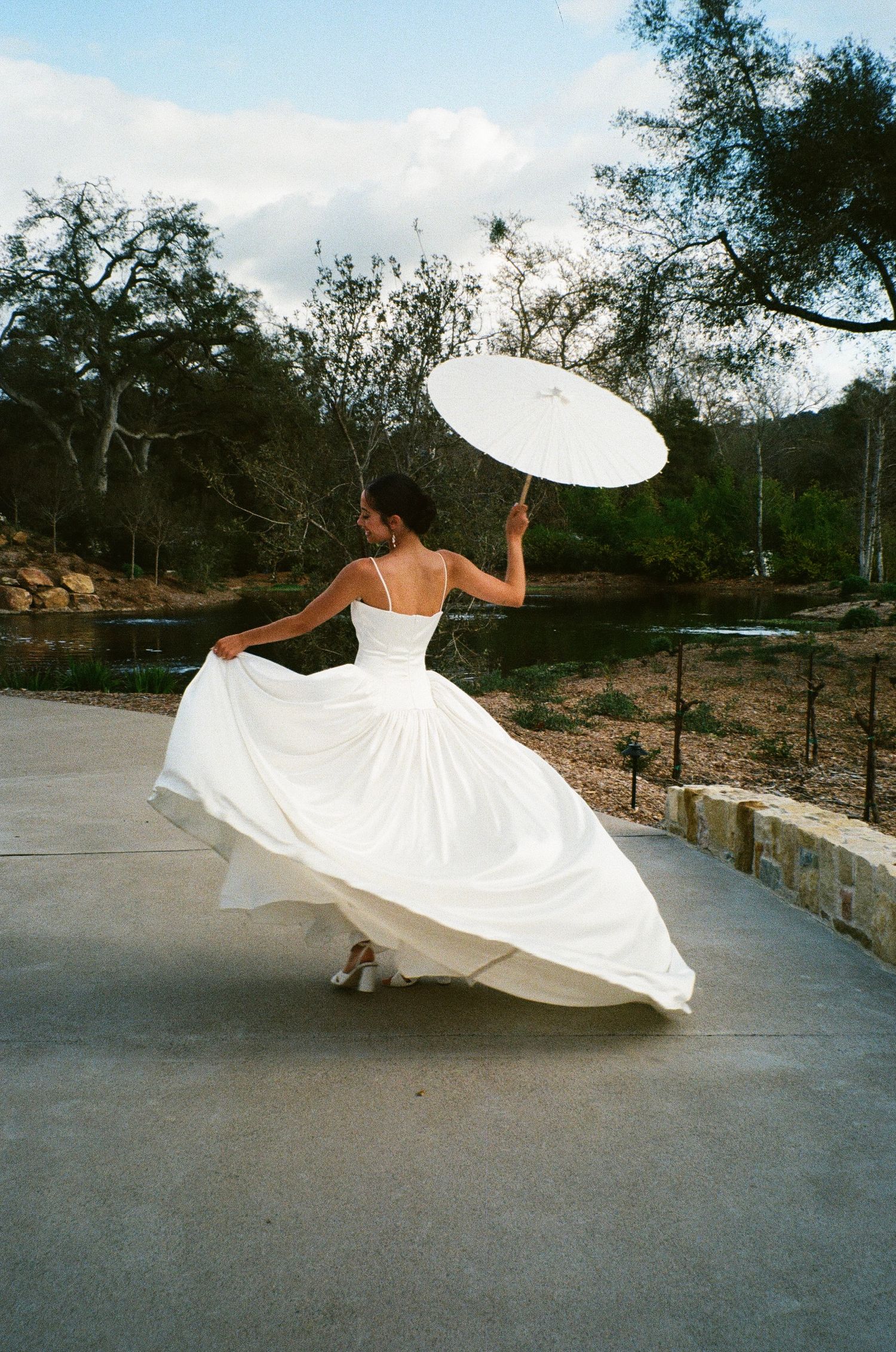 A dancer in a flowing white dress twirls gracefully on a concrete patio while holding a white parasol against a natural backdrop.