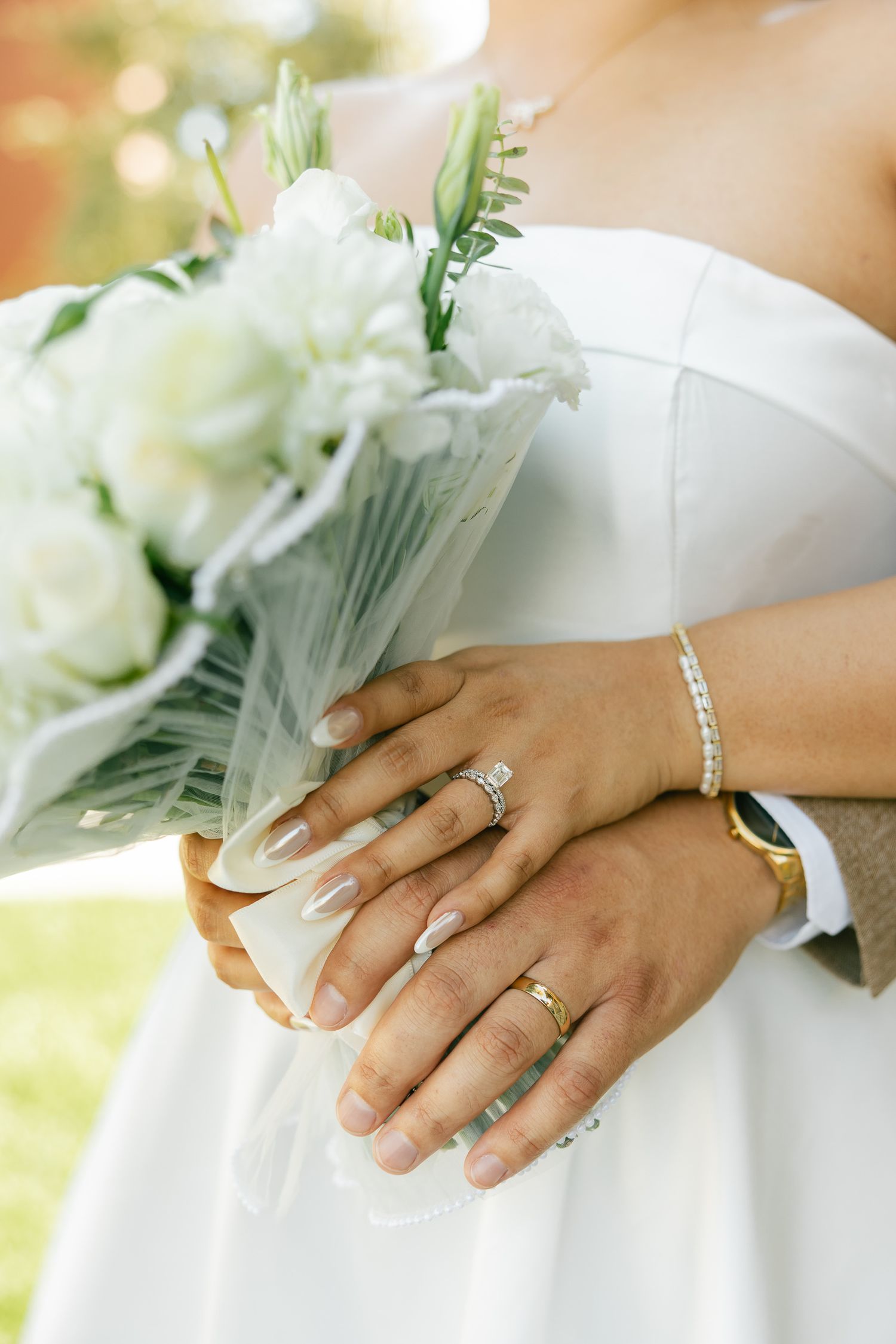 Close-up of hands with wedding rings and bracelets holding white bouquet against white dress.