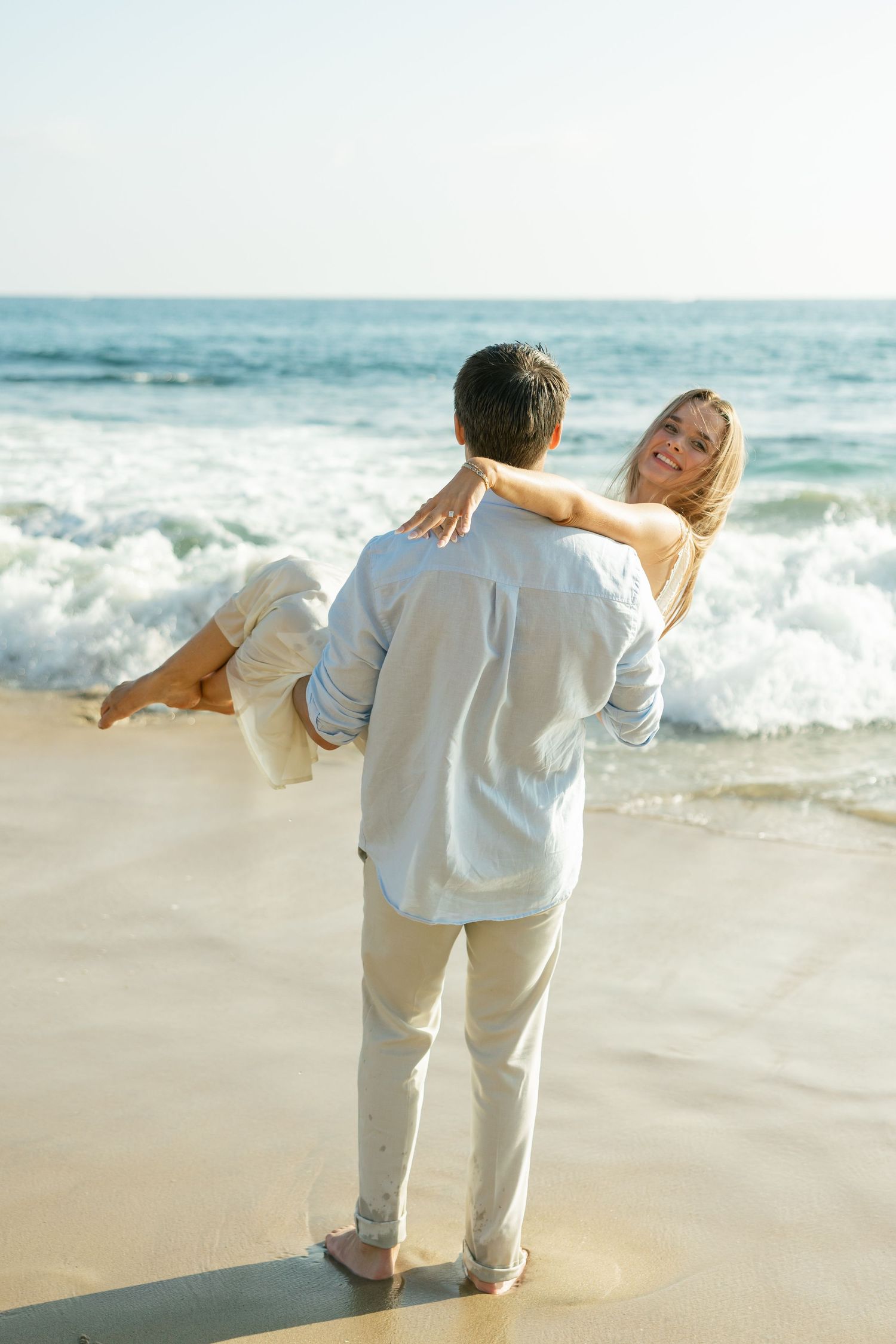 A couple in white outfits embraces on a sunny beach with crashing ocean waves in the background.