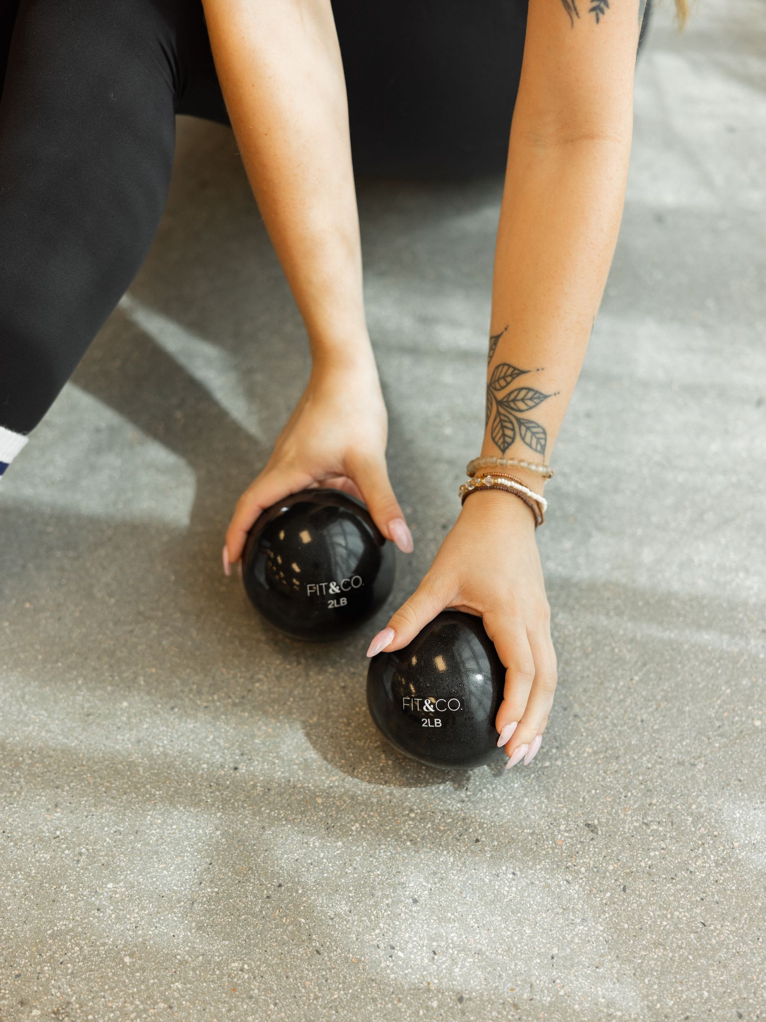 Hands with floral tattoos hold two black massage therapy balls on a concrete floor.